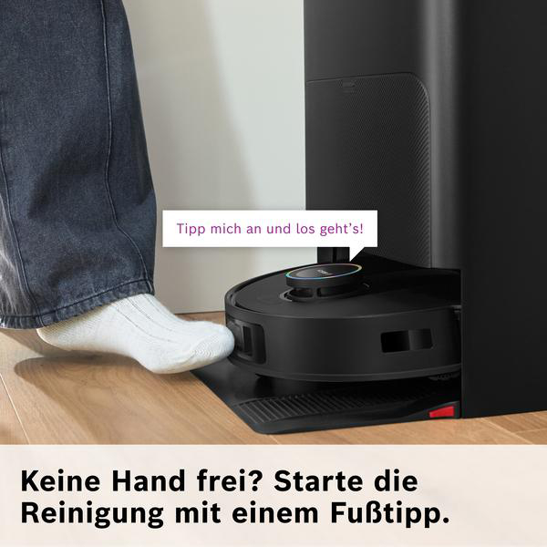 Ein Fuß tippt auf einen Saugroboter, der sich in seiner Ladestation befindet.