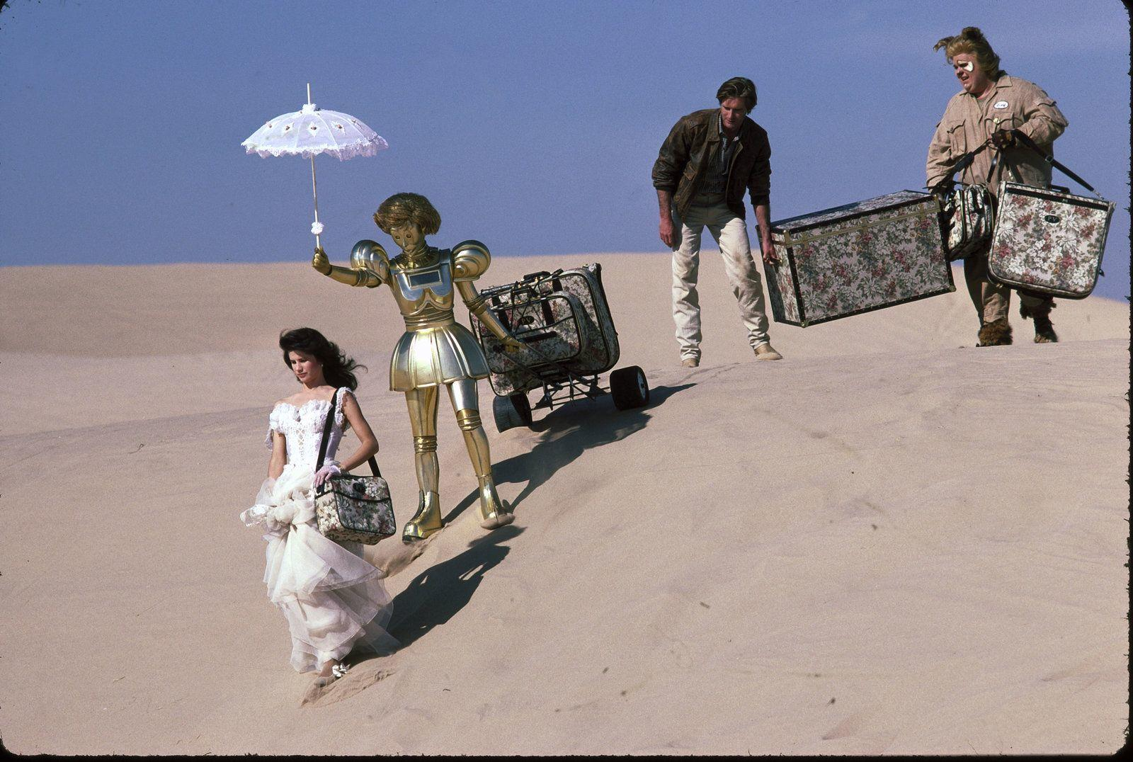 Eine Frau in einem weißen Kleid, ein goldener Roboter mit Schirm und zwei Männer mit Koffern in der Wüste.