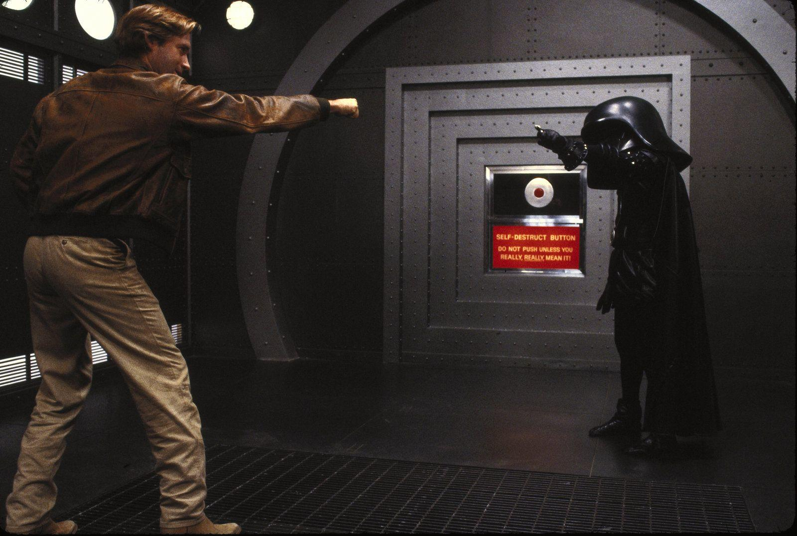 Mann in brauner Jacke steht Darth Vader gegenüber. Selbstzerstörungsknopf mit Warnung.