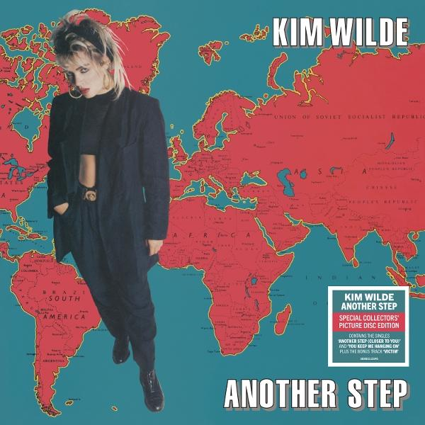 Kim Wilde steht vor einer Weltkarte. Sie trägt einen schwarzen Anzug.