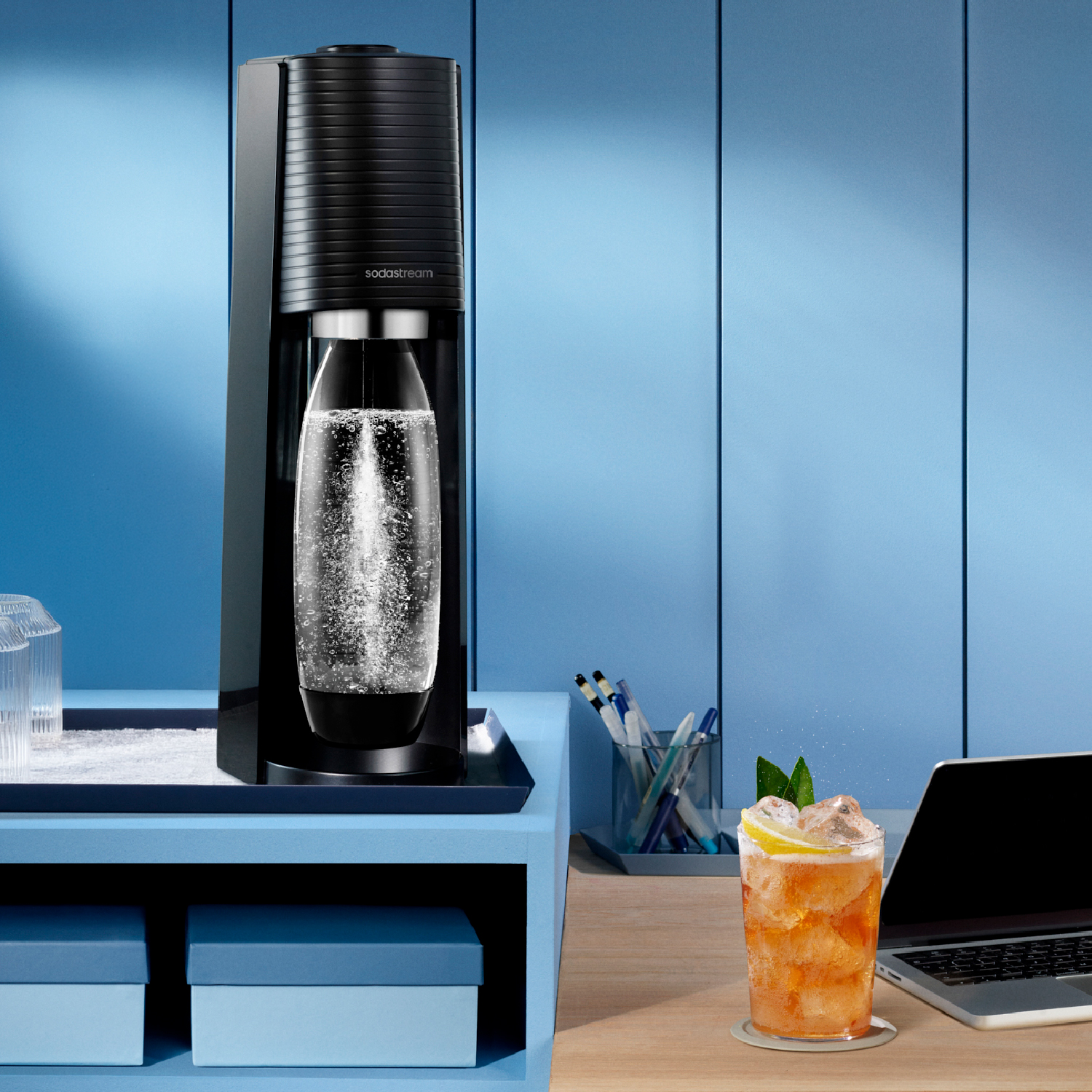 Una máquina SodaStream en un estante azul junto a una bebida y un portátil.