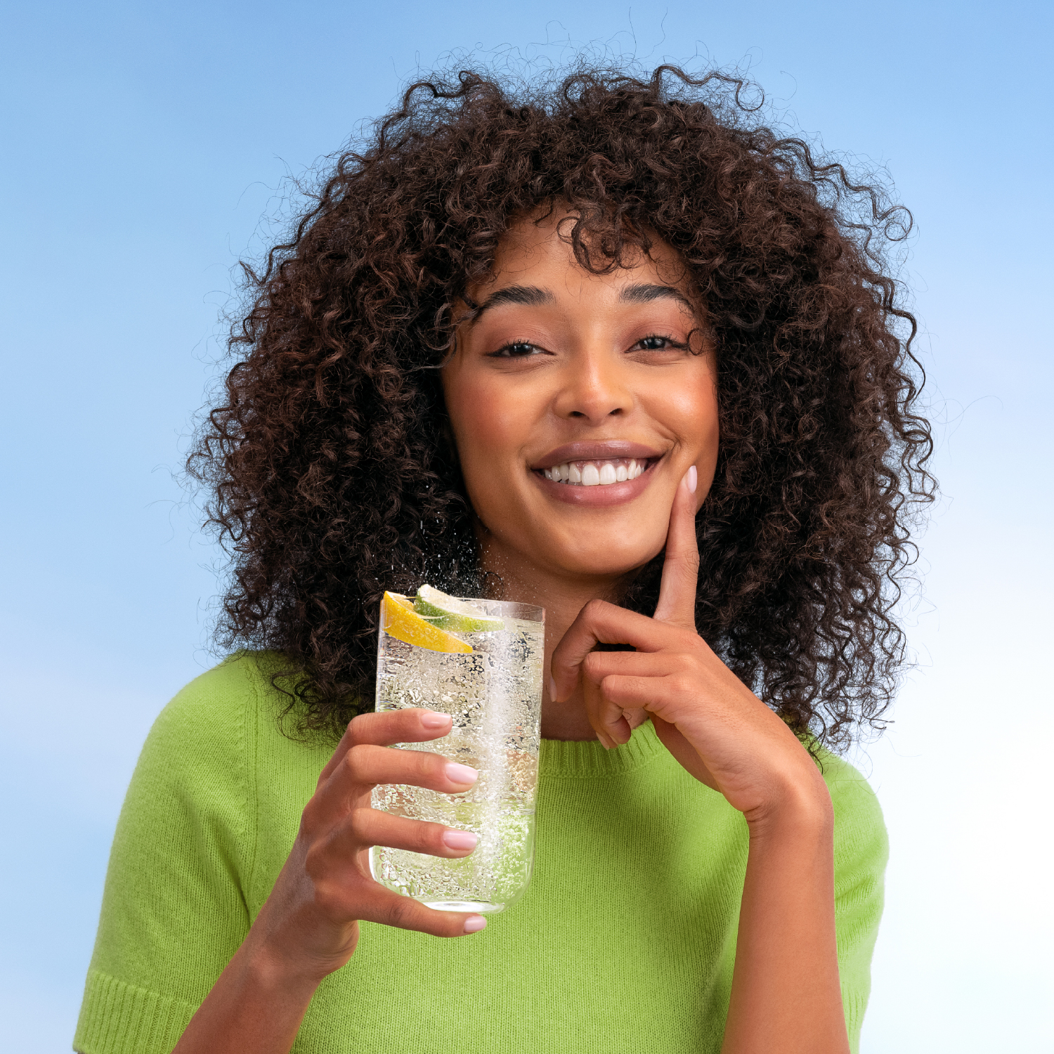 Mujer con pelo rizado sostiene un vaso con limón y lima en un suéter verde claro.