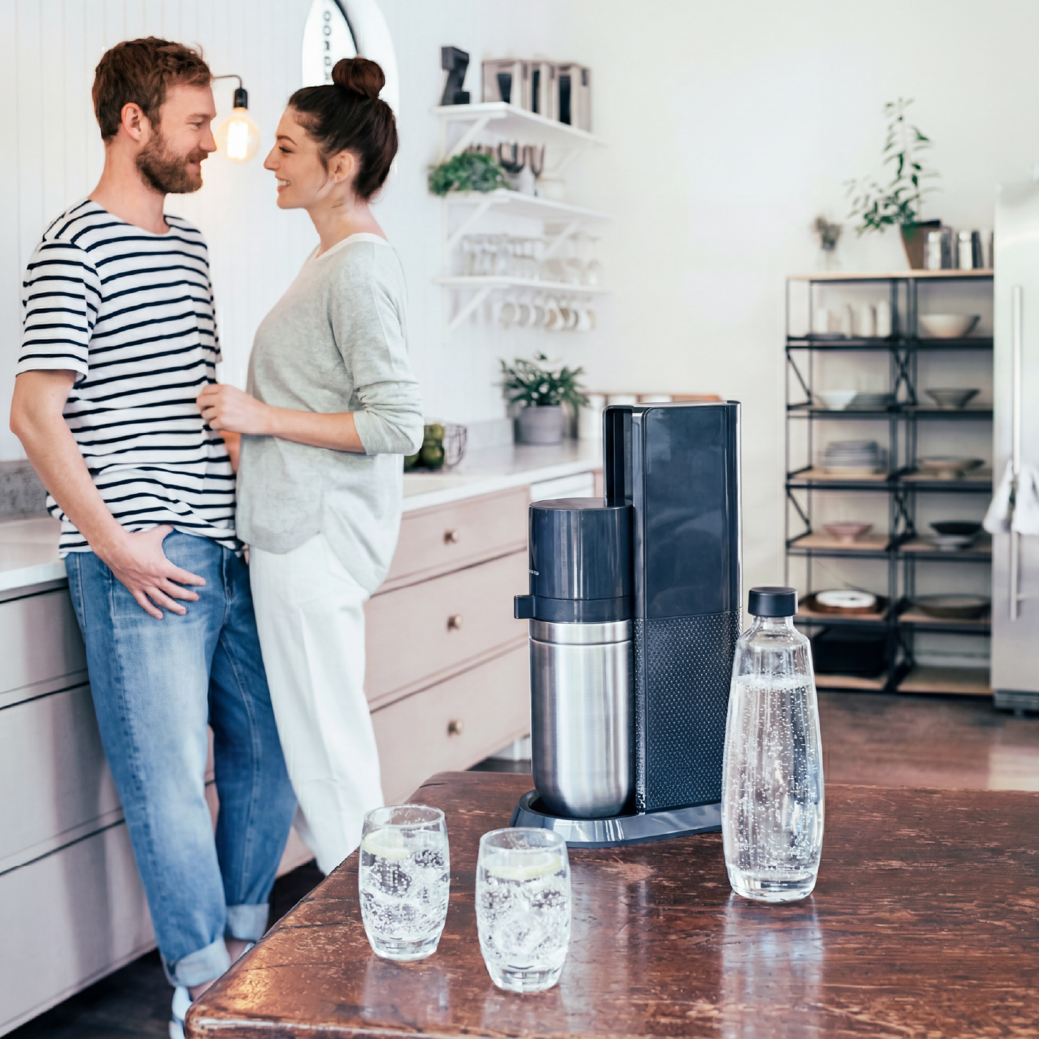 Una pareja se mira en una cocina. Hay un sifón de soda en la mesa.