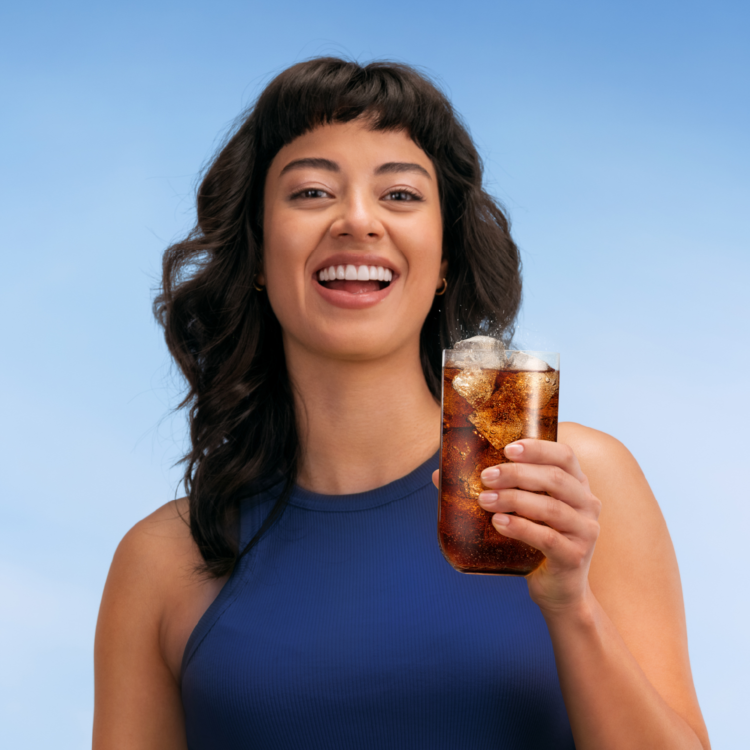 Mujer sosteniendo un vaso de cola con hielo contra un cielo azul claro.