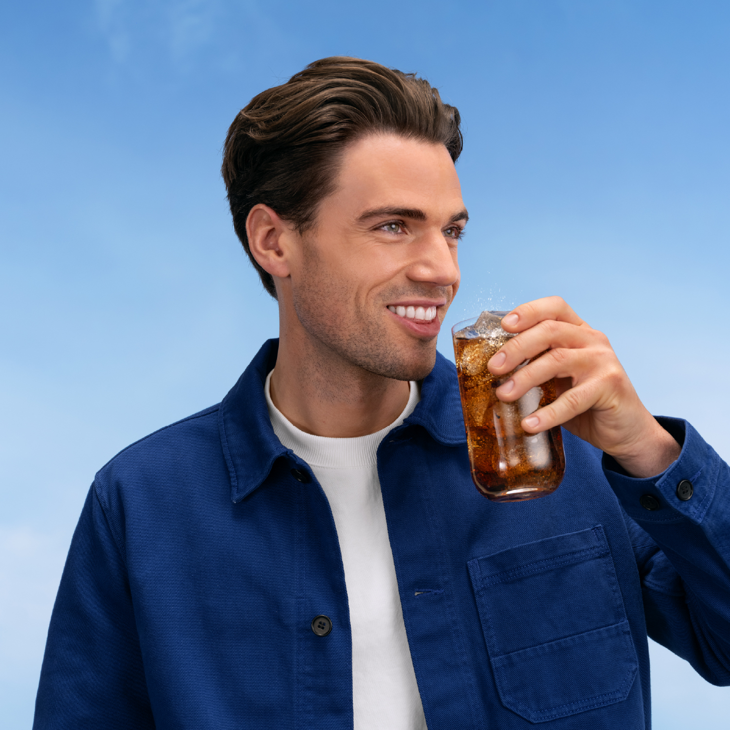 Un hombre bebe un vaso de cola con hielo y salpicaduras contra el cielo azul.