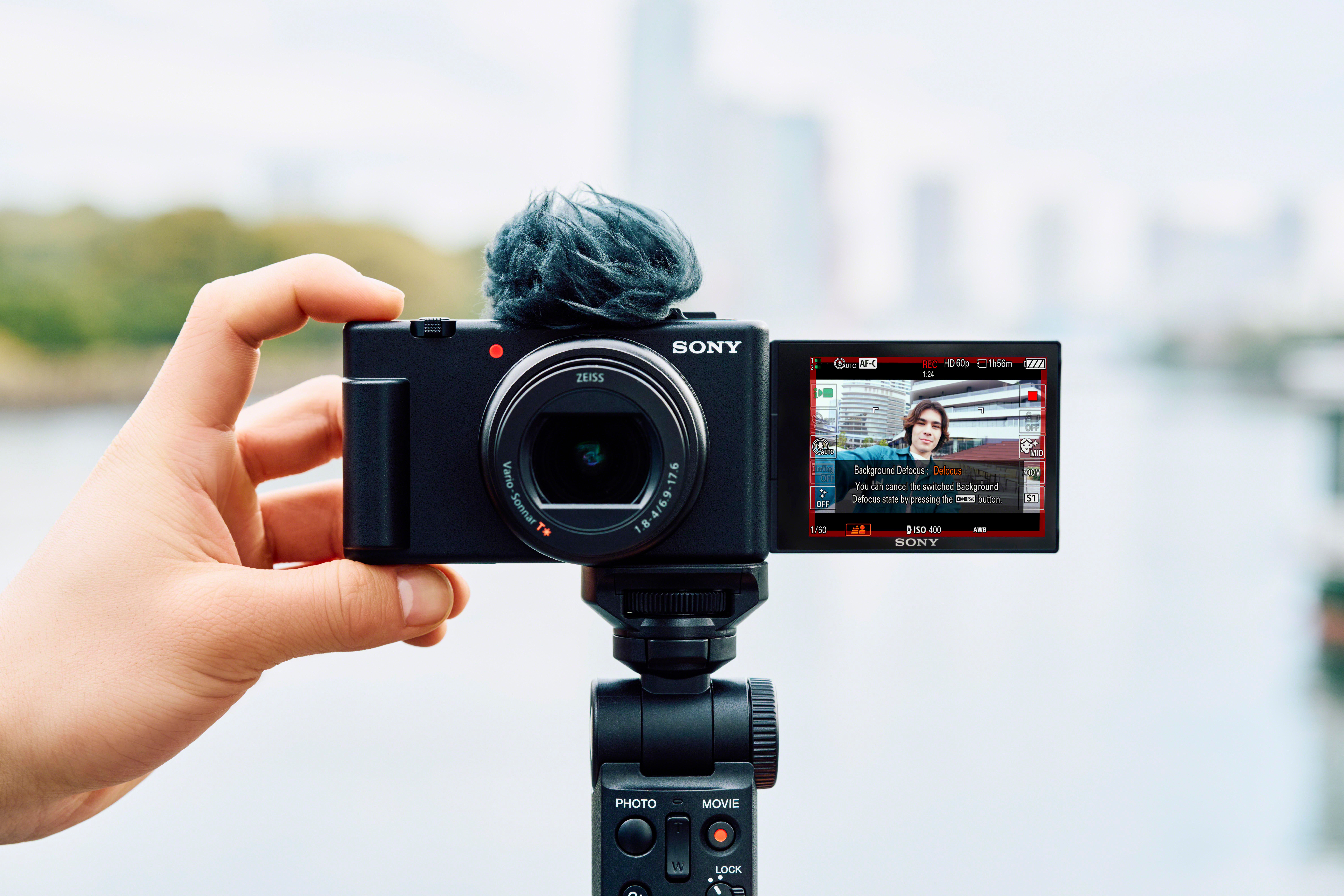 Hand hält eine Sony Vlogging-Kamera mit flauschigem Mikrofon. Bildschirm zeigt einen Mann.