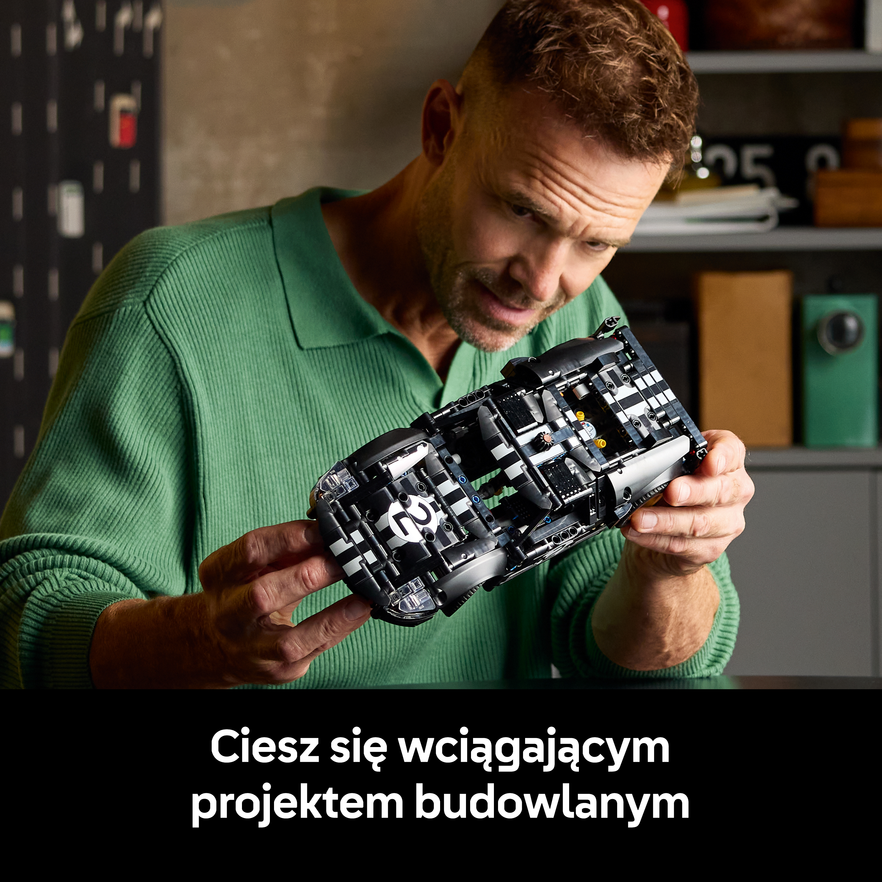 Mężczyzna w zielonym swetrze trzyma czarny model samochodu w kratkę.