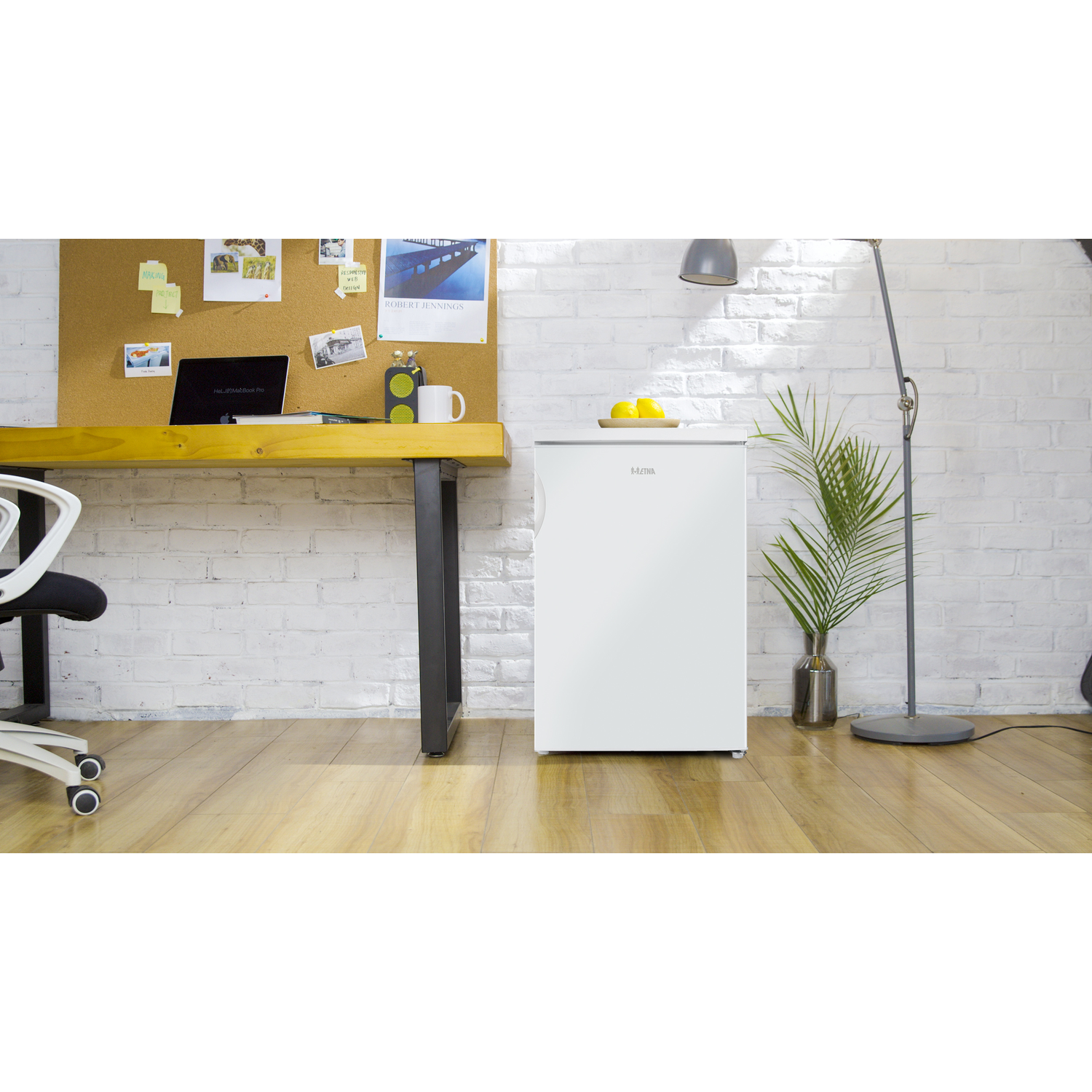 Een witte mini-koelkast op houten vloer naast een bureau met prikbord.