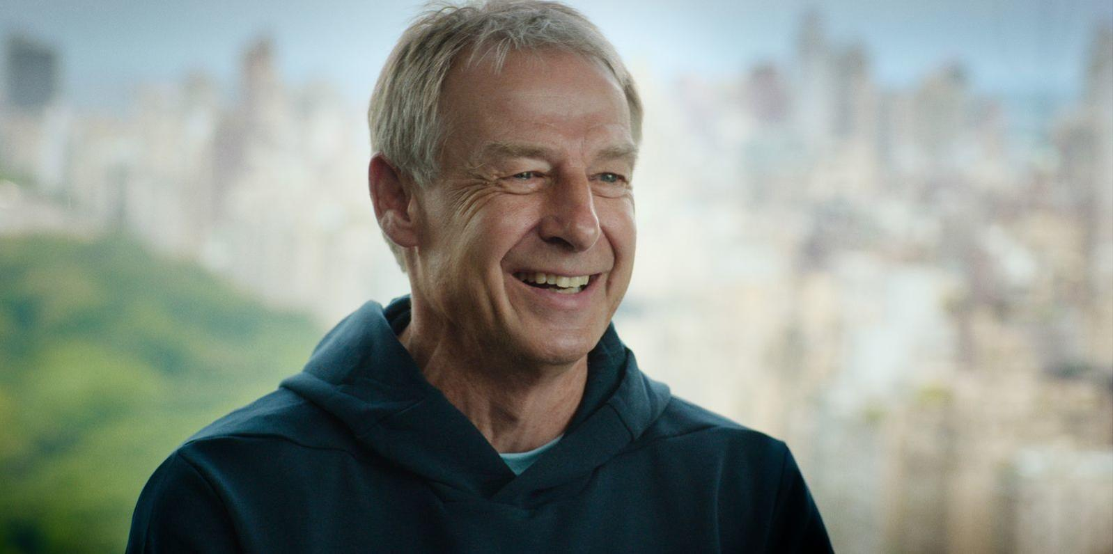 Ein Mann mittleren Alters mit grauem Haar lacht, trägt einen dunkelblauen Hoodie.