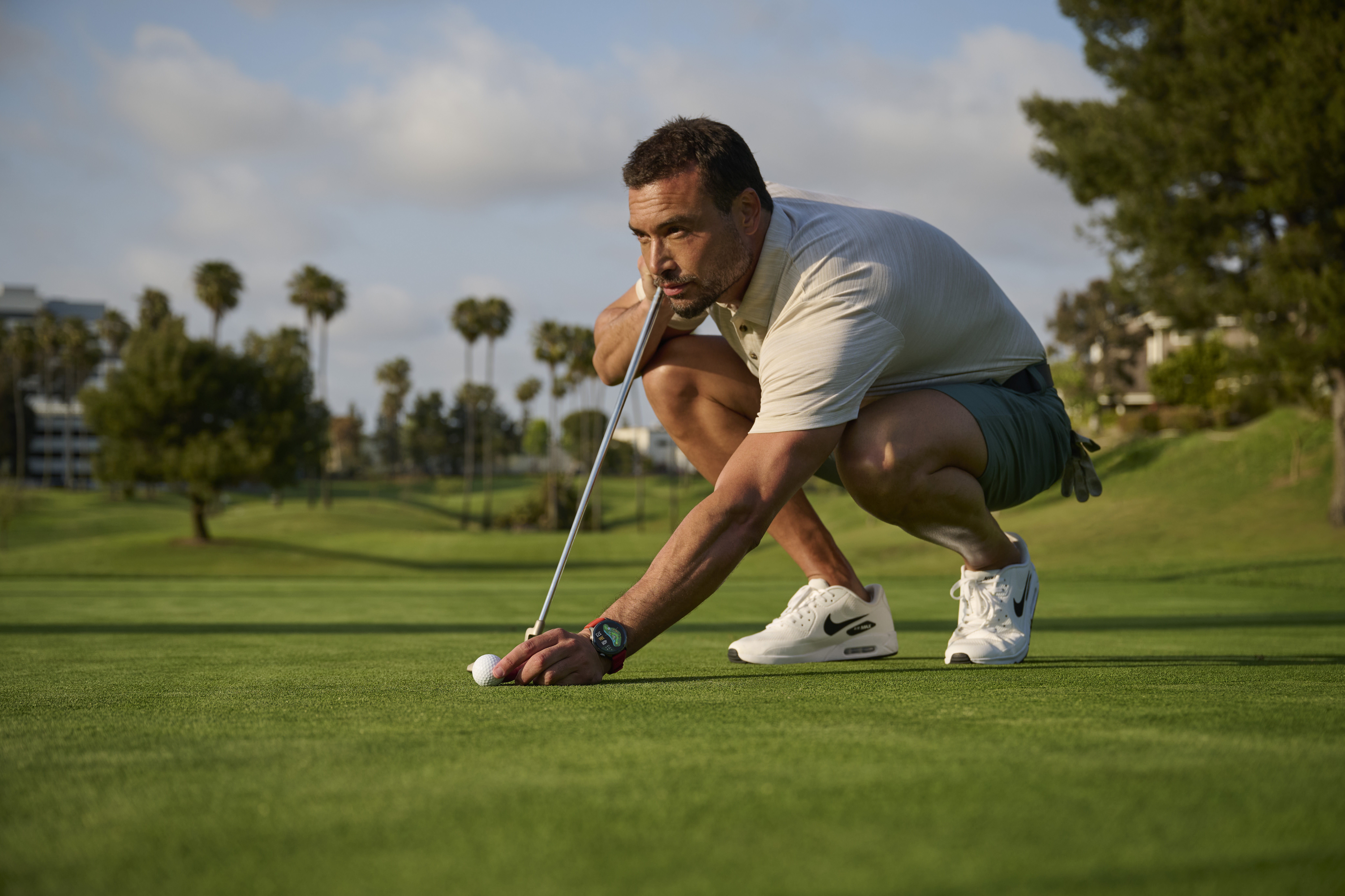 Golfista klęczy na zielonym polu golfowym, zaznaczając piłkę kijem.