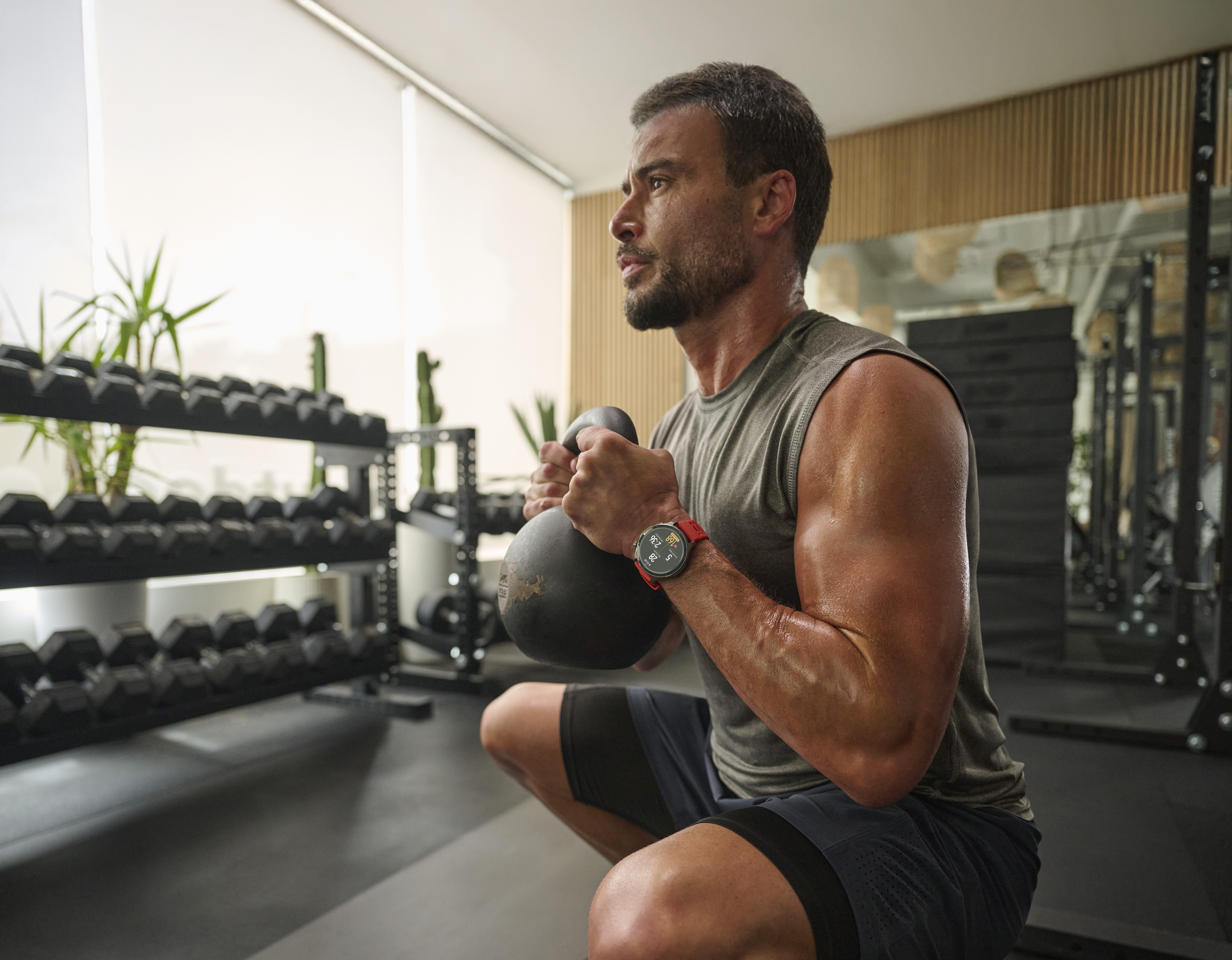 Mężczyzna robi przysiady z kettlebell w siłowni, stojaki na hantle w tle.