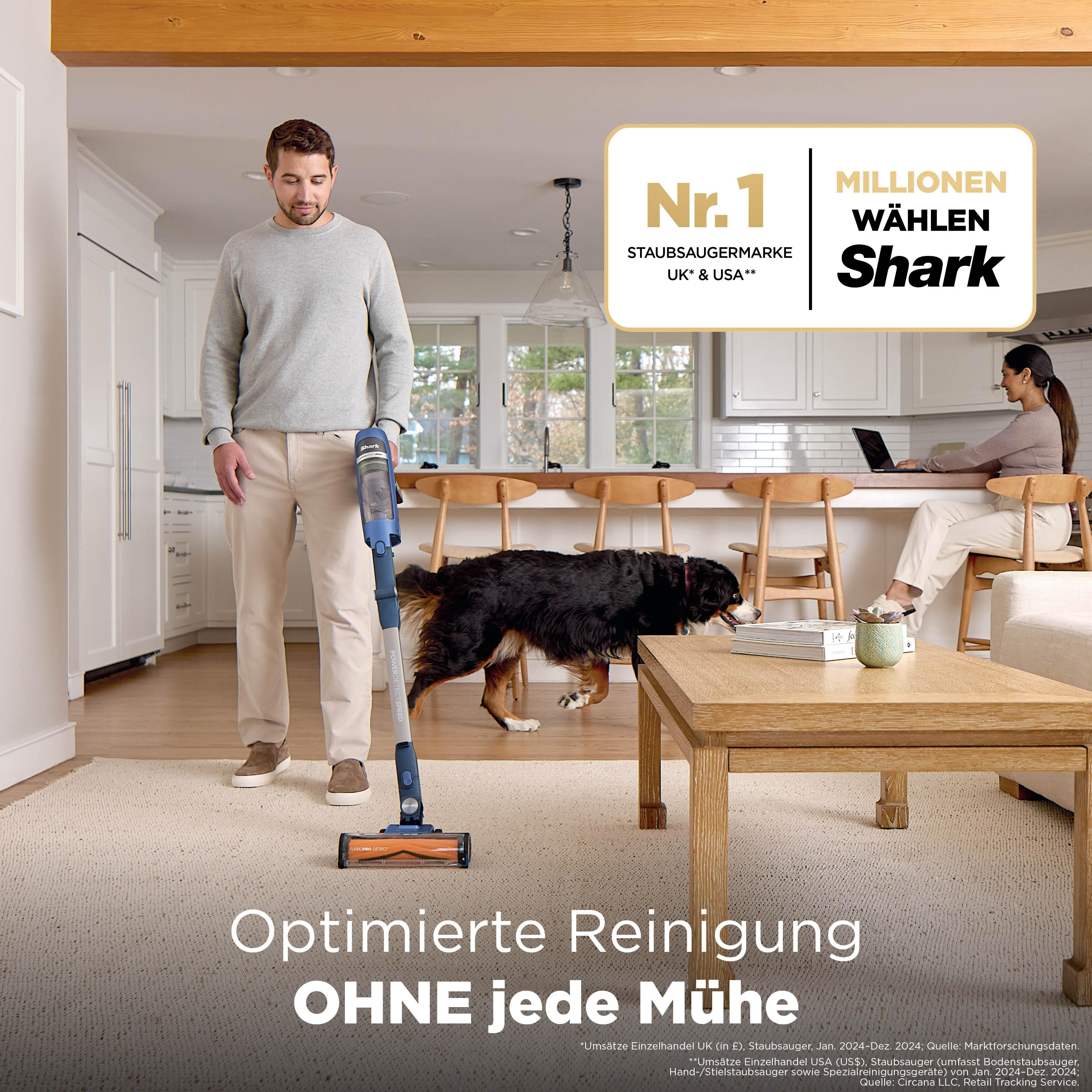 Mann mit Staubsauger im Haus, Hund und Frau am Laptop im Hintergrund.