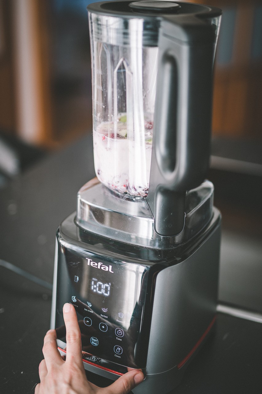Egy kéz megnyomja a 'smoothie' gombot egy Tefal turmixgépen, gyümölccsel és folyadékkal.