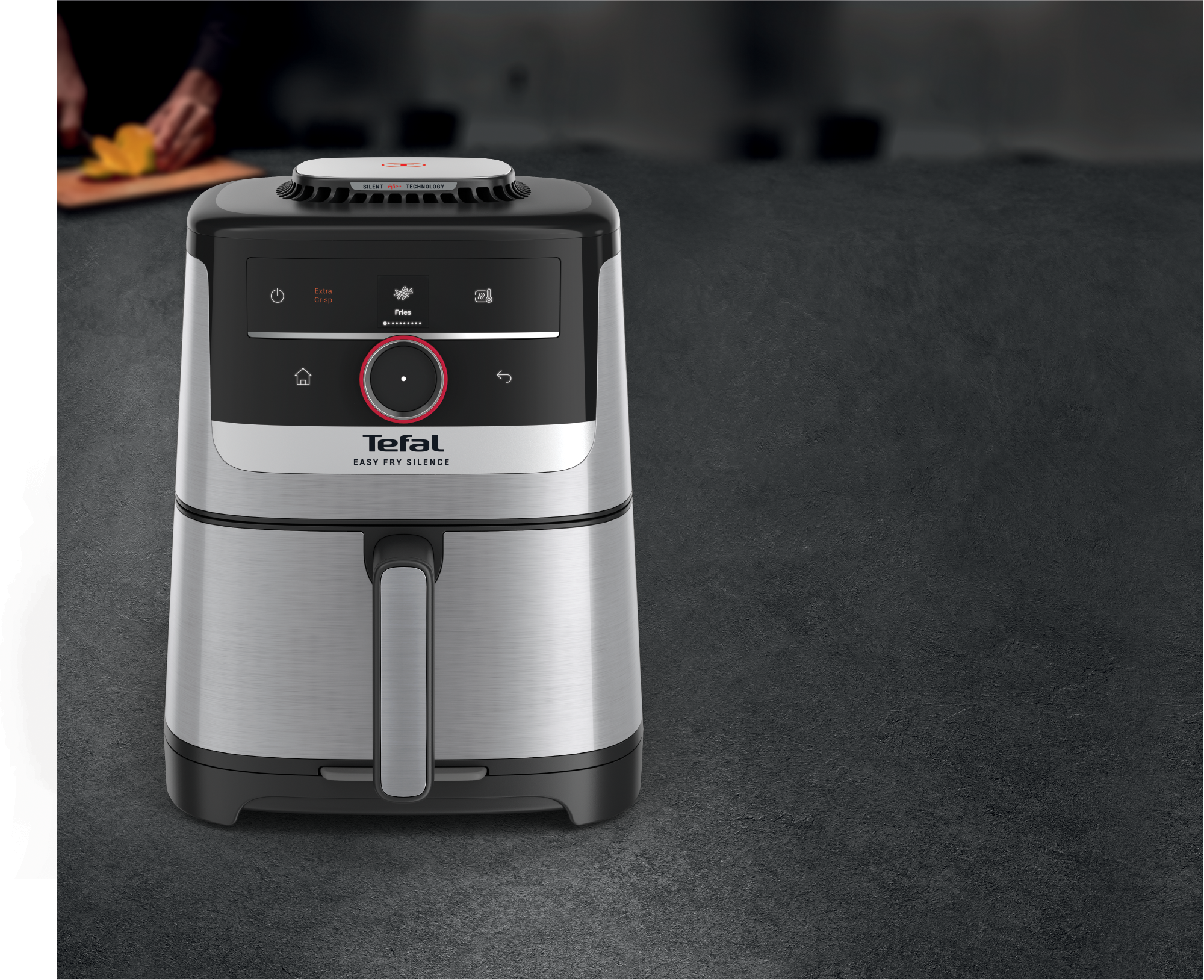 Ezüst és fekete Tefal légsütő áll sötét felületen. Egy személy gyümölcsöt vág a háttérben.