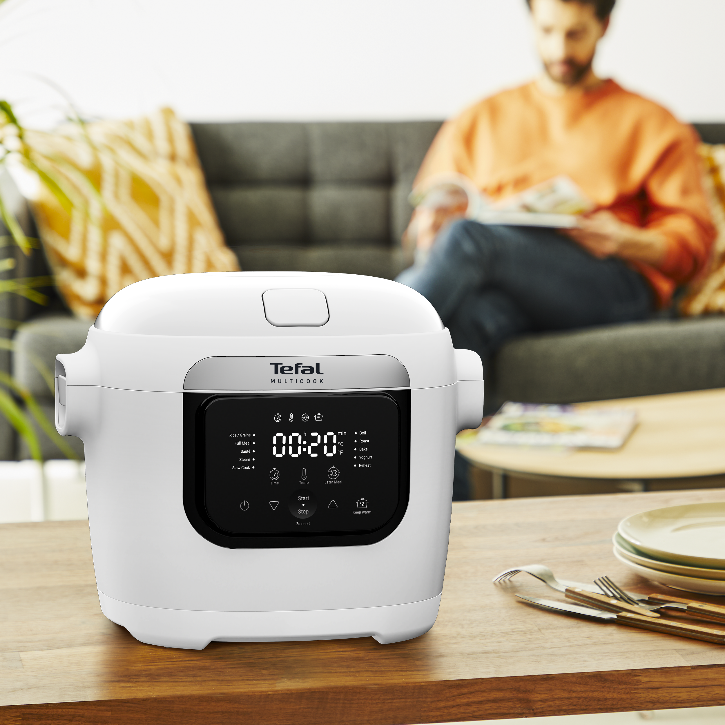 Egy fehér Tefal multifunkciós főző egy faasztalon. A háttérben egy férfi olvas.