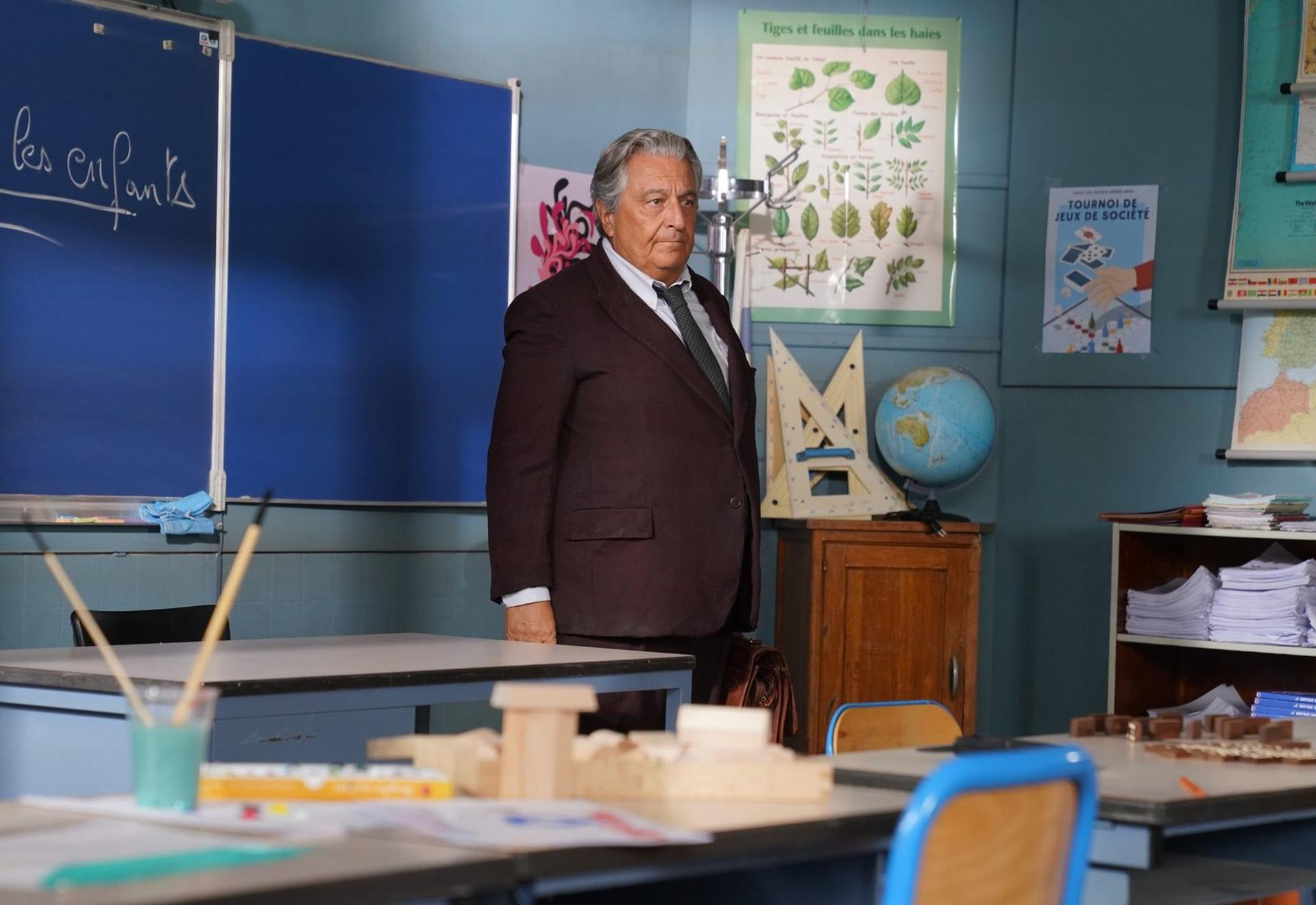 Ein älterer Mann im Anzug steht in einem Klassenzimmer mit einer blauen Tafel und Postern.