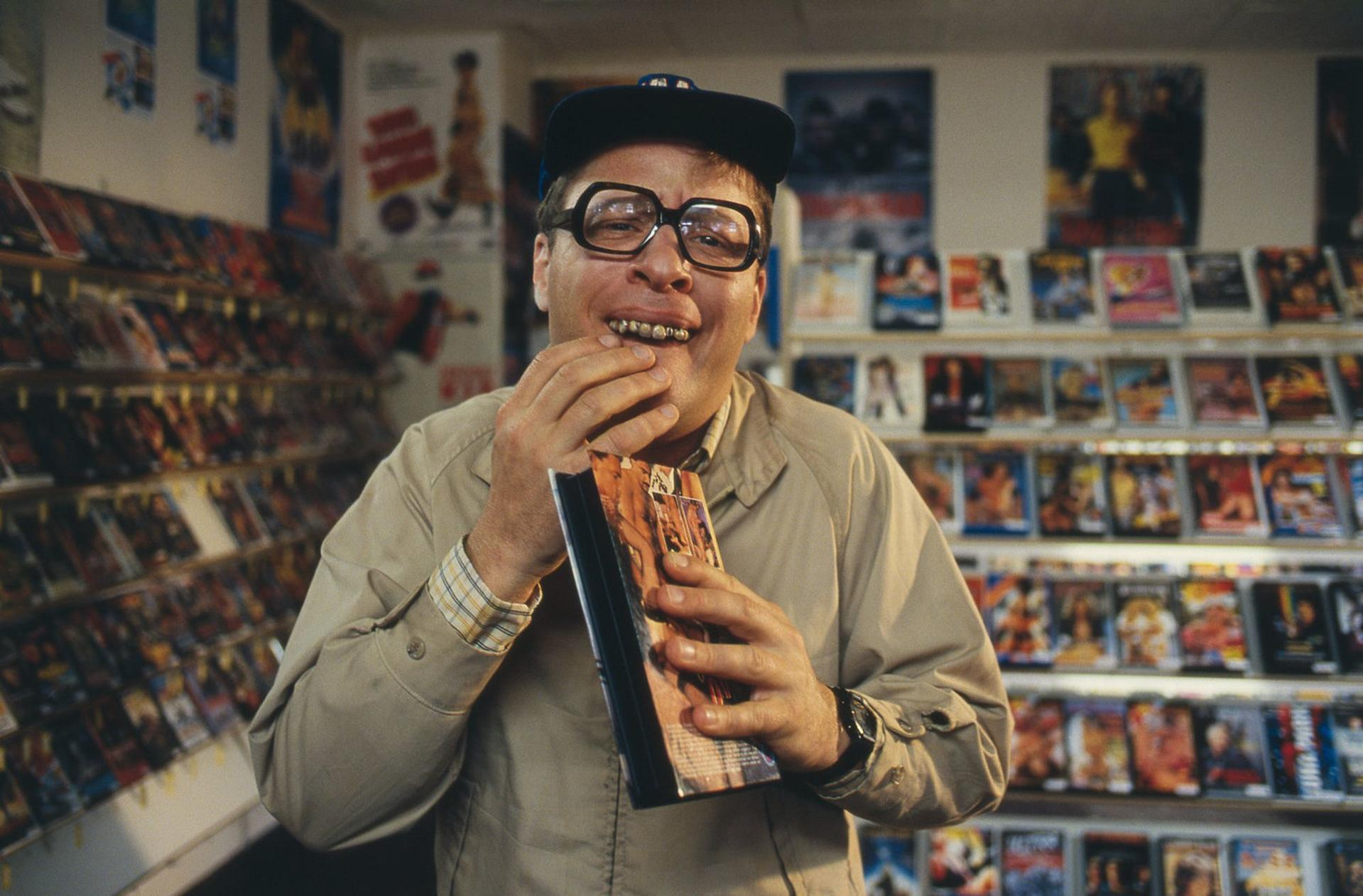 Ein Mann mit Brille und Kappe hält eine Videokassette in einem Laden mit Regalen voller Kassetten.