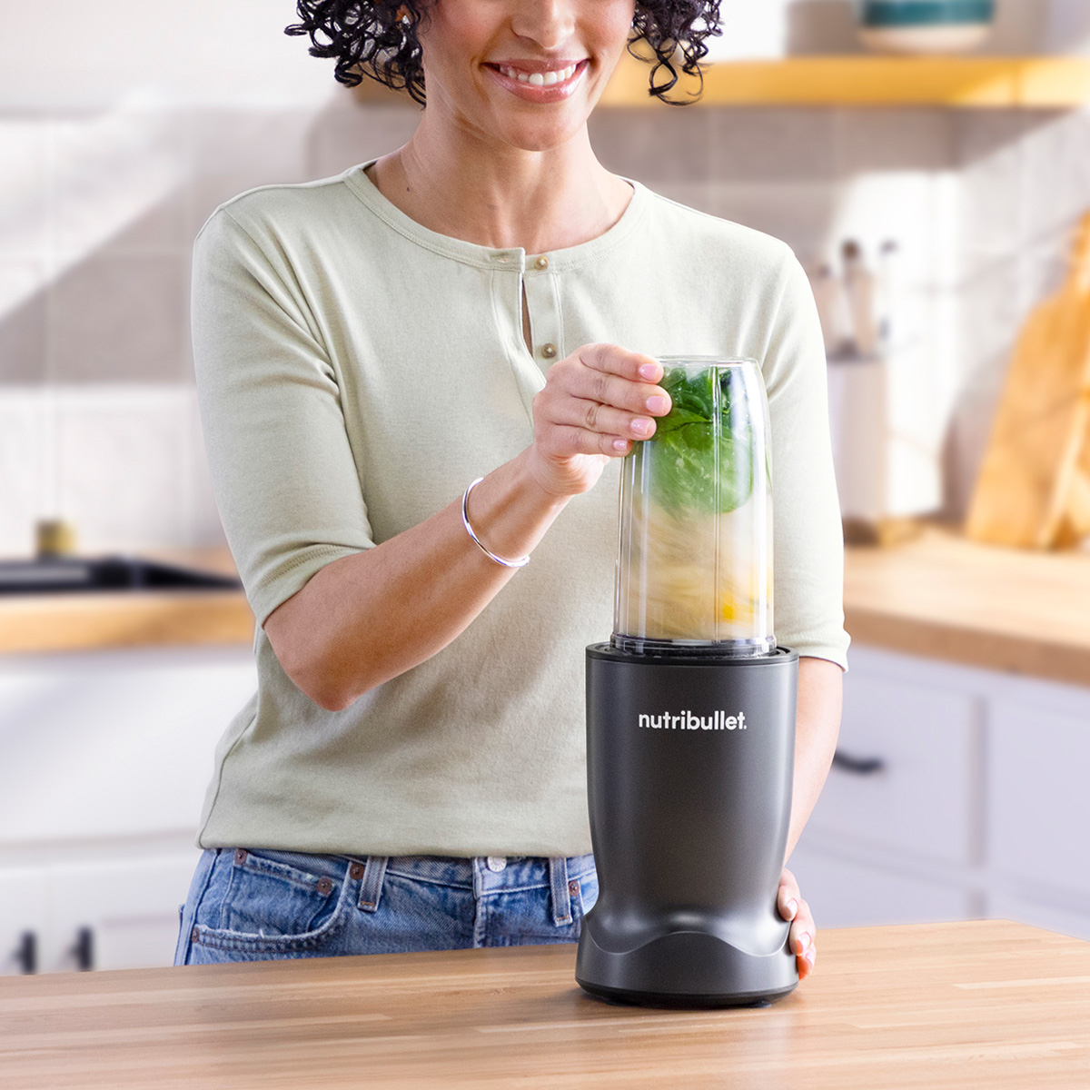 Frau mit Locken mixt grünes Blattgemüse und gelbe Fruchtstücke in einem Mixer.