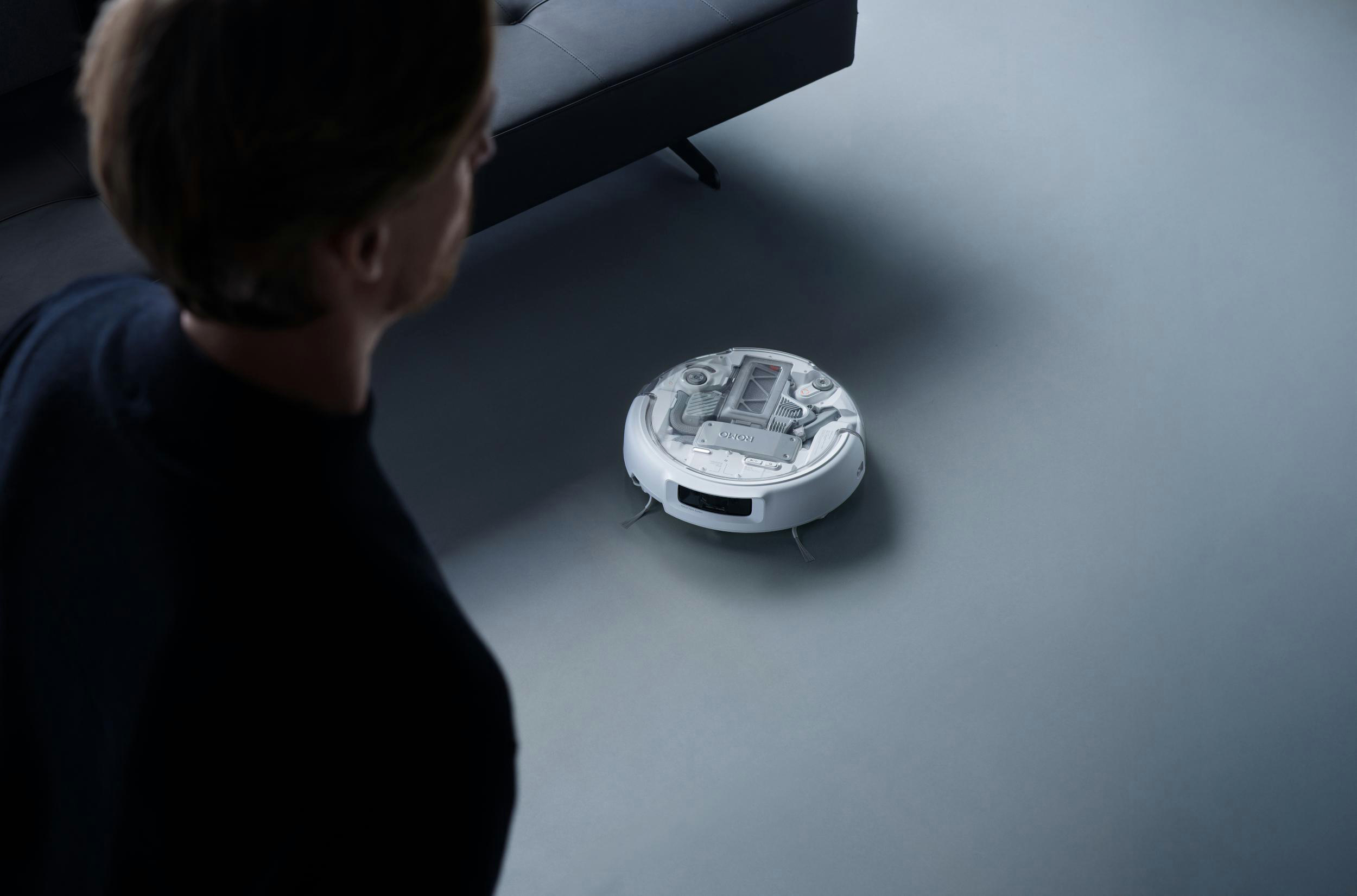 Ein Mann blickt auf einen weißen Saugroboter auf einem grauen Boden neben einem Sofa.