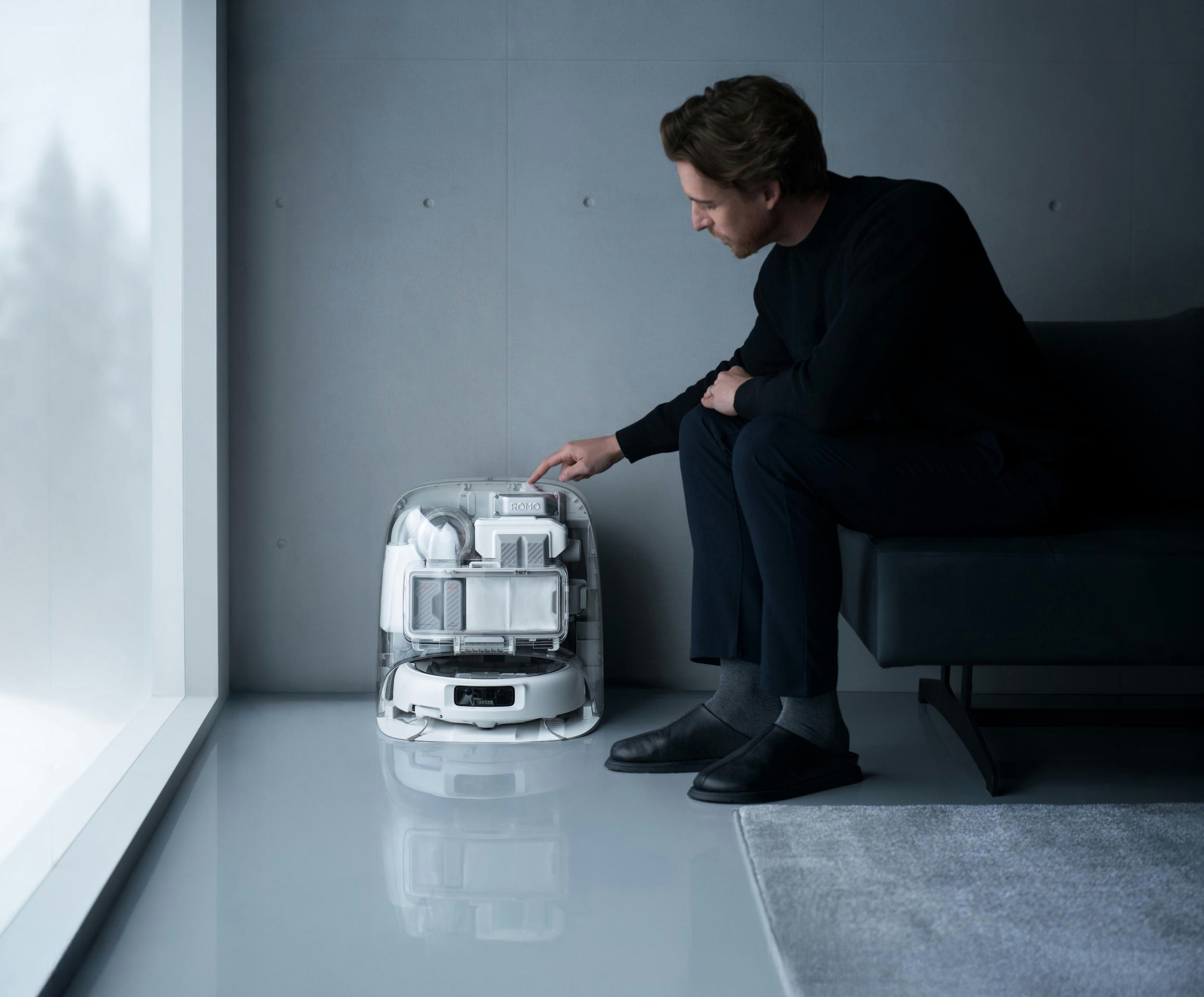Ein Mann berührt einen transparenten Roboter-Staubsauger. Innen sind weiße Behälter zu sehen.