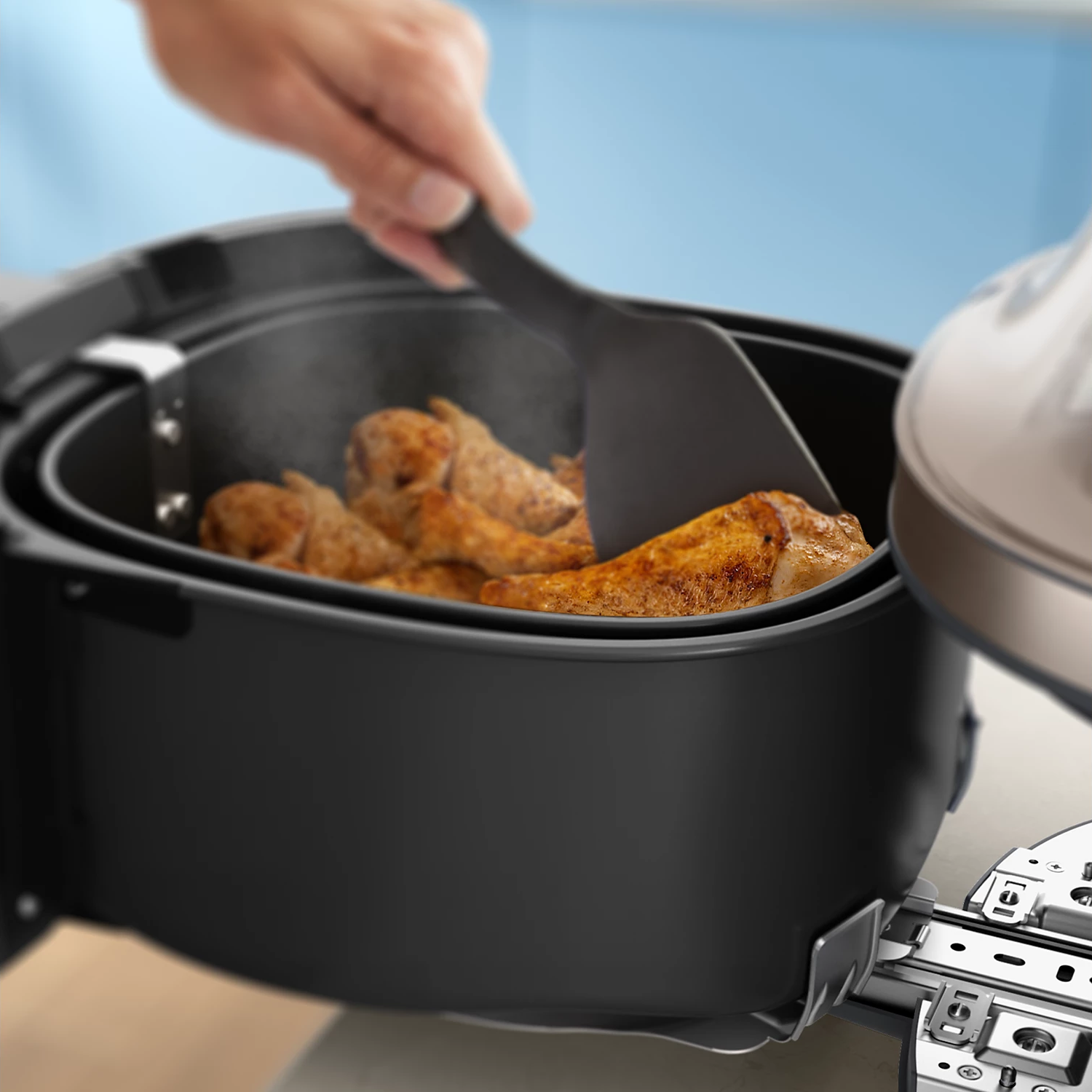 Ein Kochwender wendet Hähnchenschenkel in einem schwarzen Heißluftfritteusenkorb.