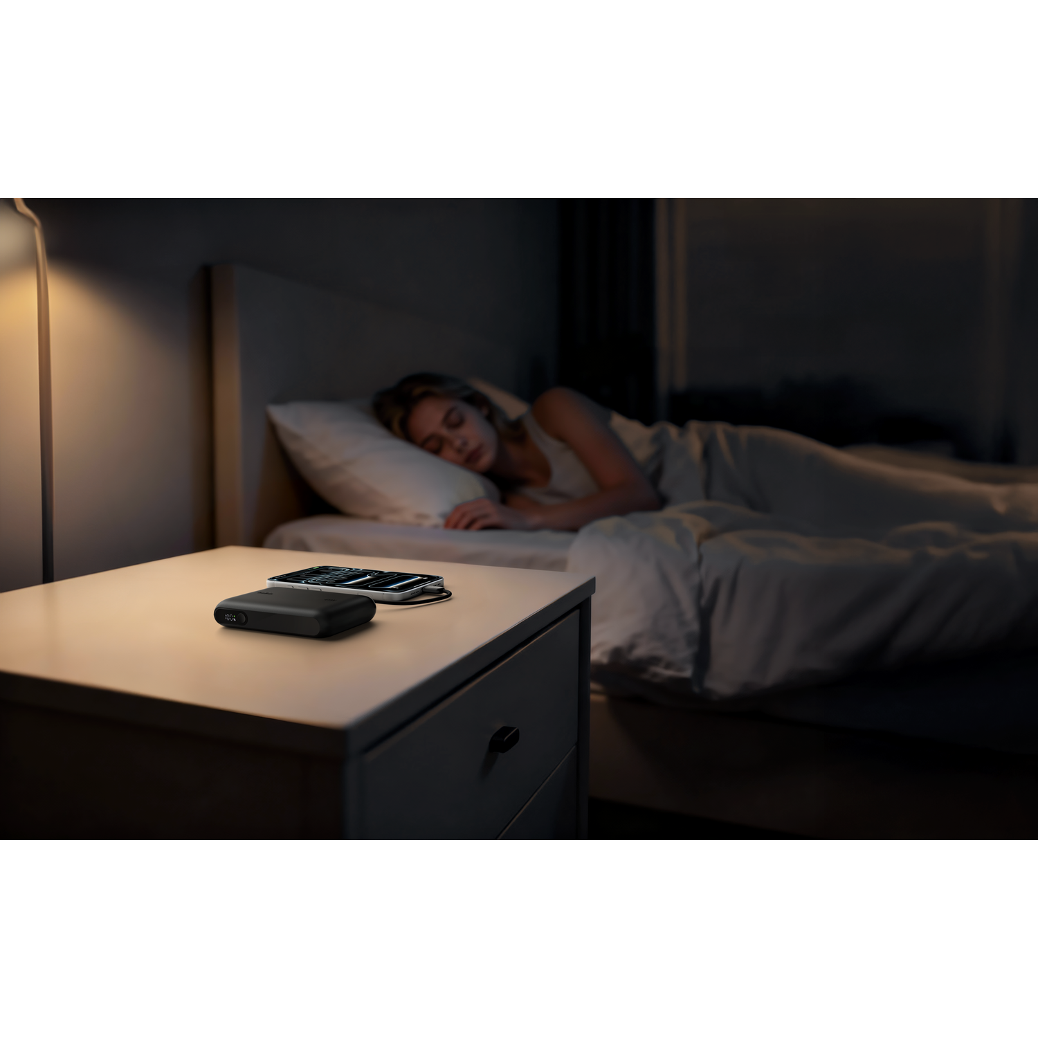Woman sleeps in bed, nightstand with charger and phone.