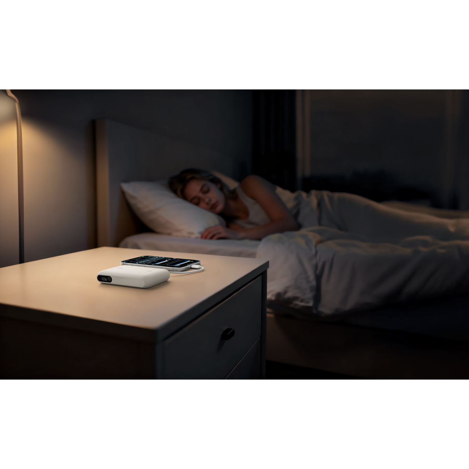 A woman sleeps in bed, nightstand with charging phone and white device.