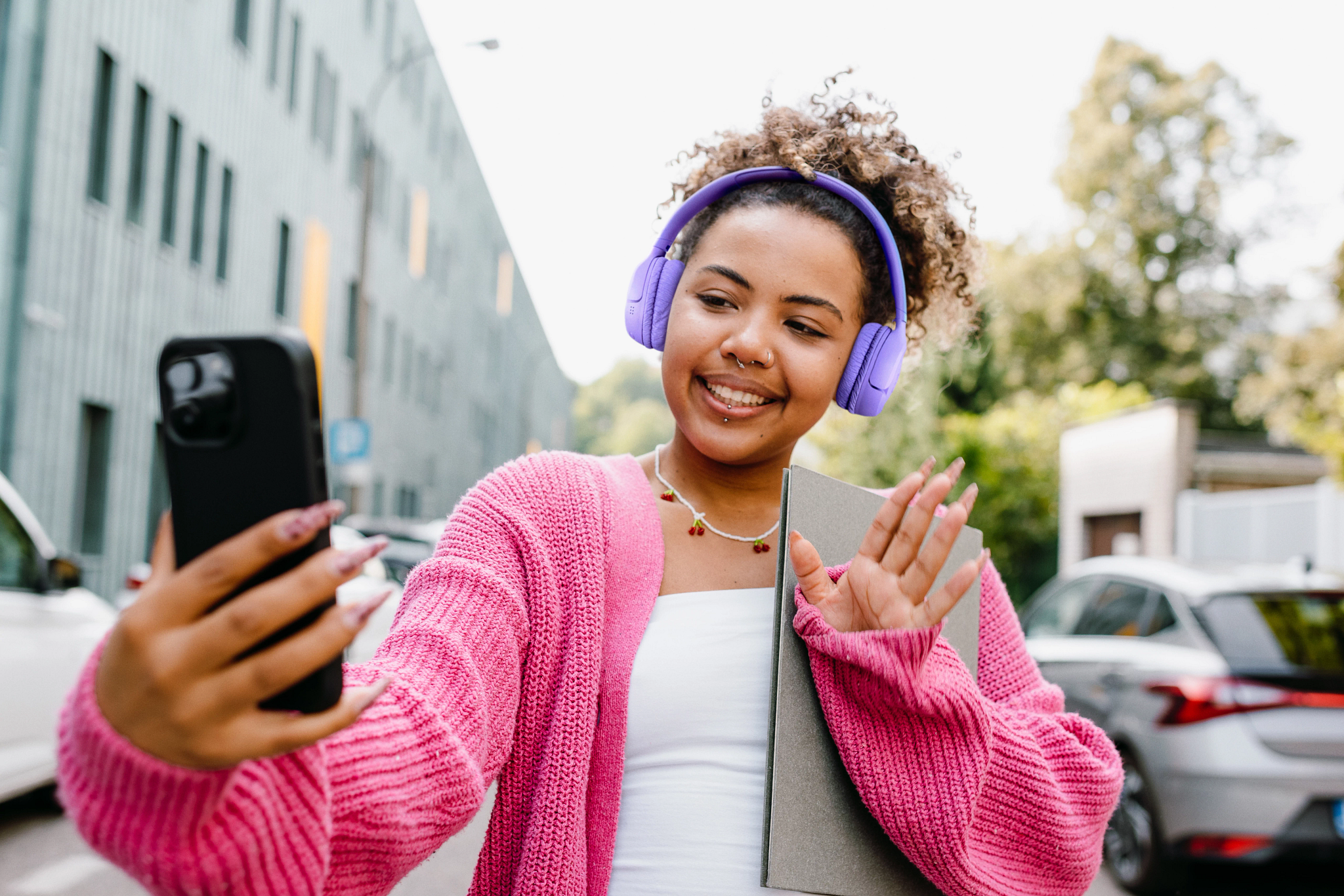 Junge Frau mit lila Kopfhörern hält Handy und winkt, trägt pinken Cardigan.