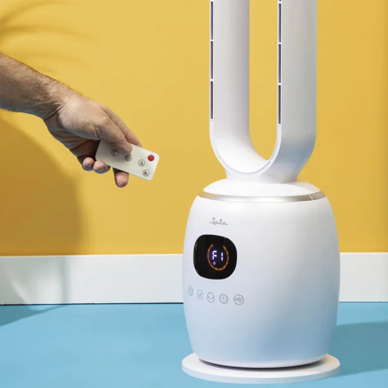 Una mano sostiene un mando a distancia hacia un ventilador de torre blanco con pantalla digital.