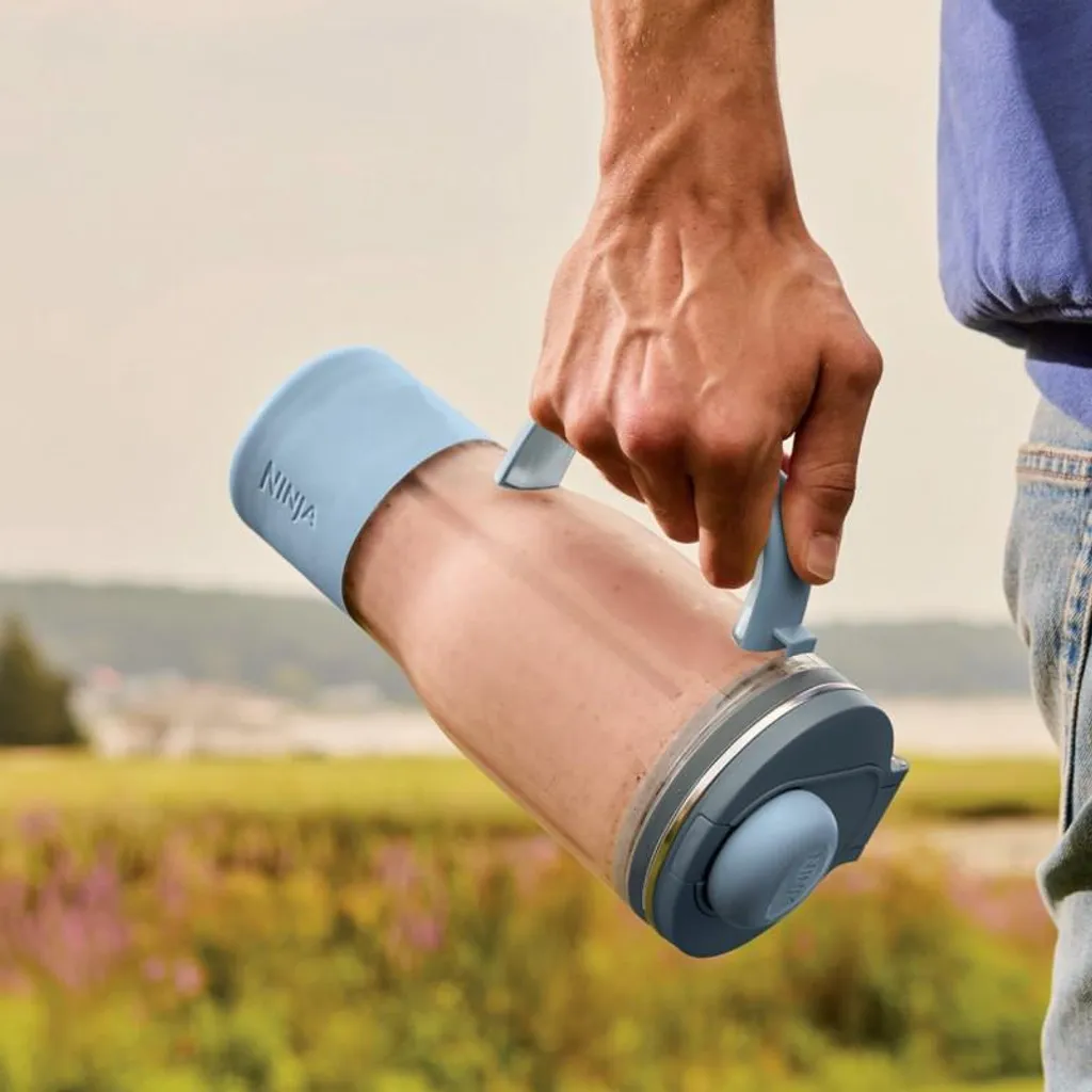 Hombre sostiene un vaso de licuadora transparente con tapa y asa azul claro.