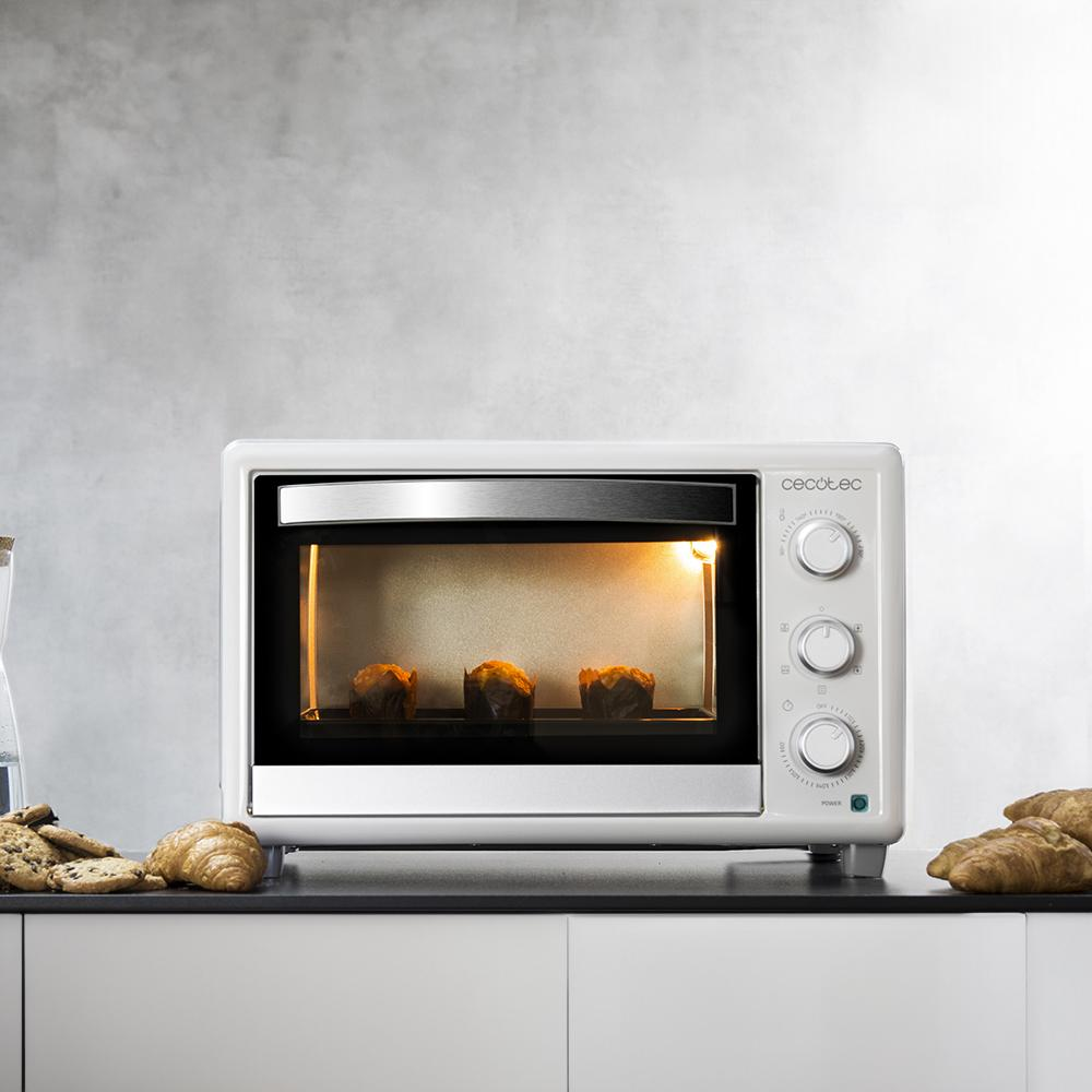 Un horno mini blanco con tres magdalenas horneándose dentro. Al lado hay cruasanes y galletas.