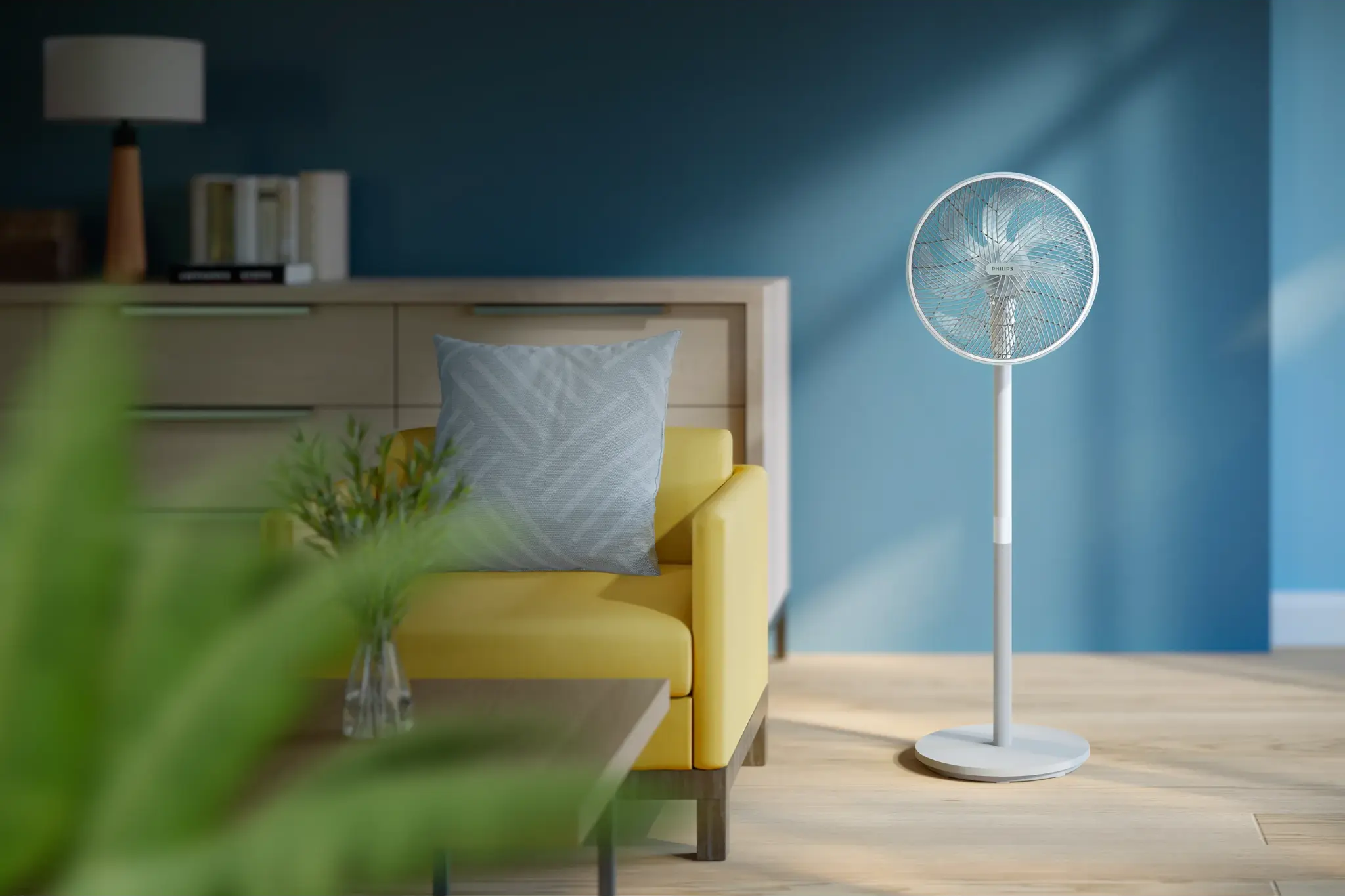 Ein Standventilator steht neben einem gelben Sofa mit blauem Kissen in einem Raum.