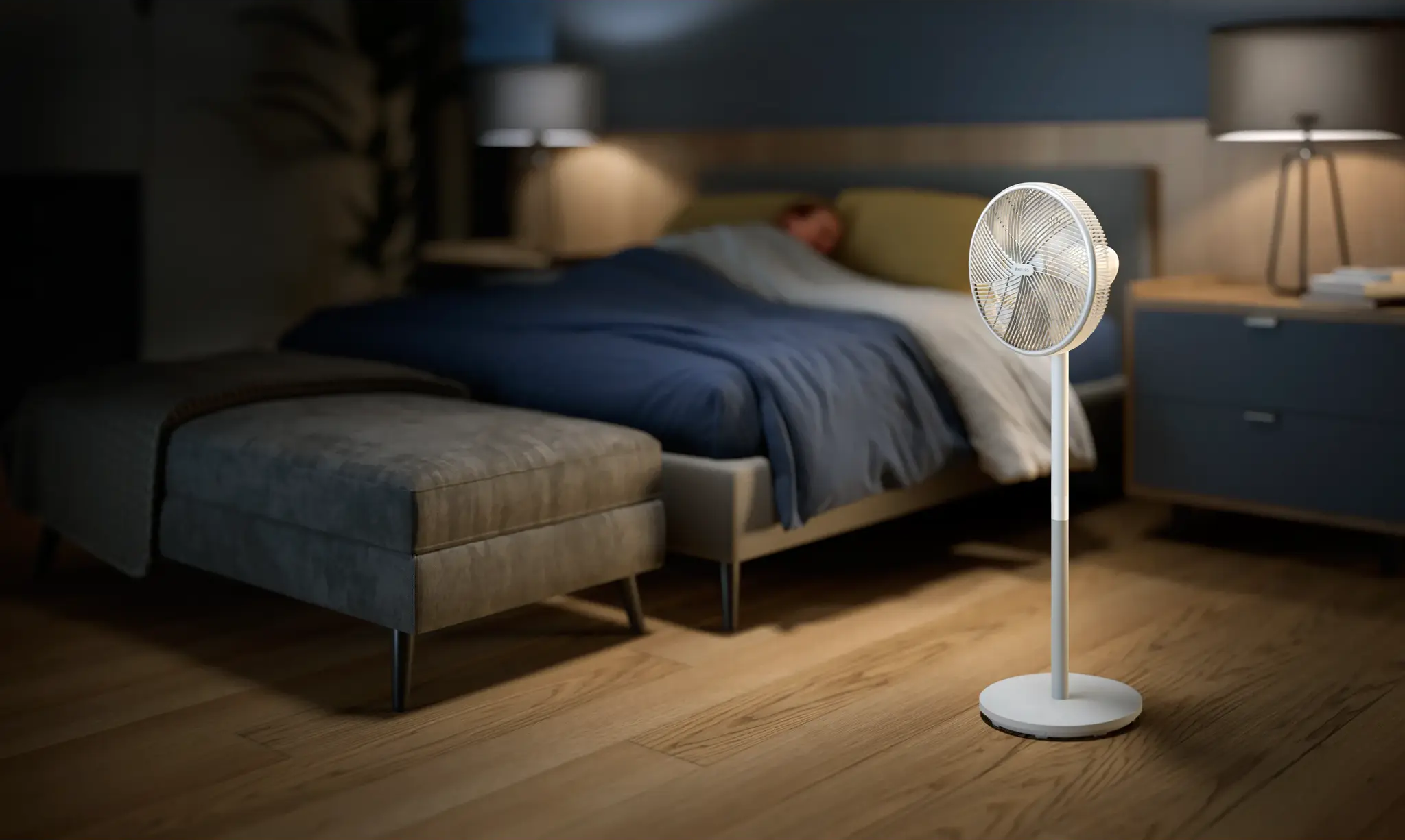 Ein weißer Standventilator steht auf einem Holzboden neben einem Bett. Eine Person schläft unter blauer Decke.