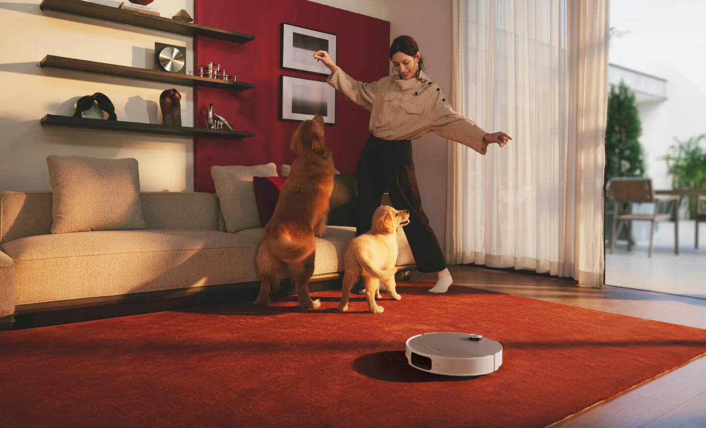 Donna gioca con due cani su tappeto rosso, robot aspirapolvere in primo piano.