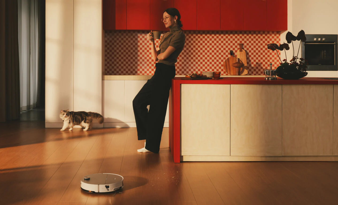 Un robot aspirapolvere pulisce un pavimento in legno in una cucina con una donna e un gatto.