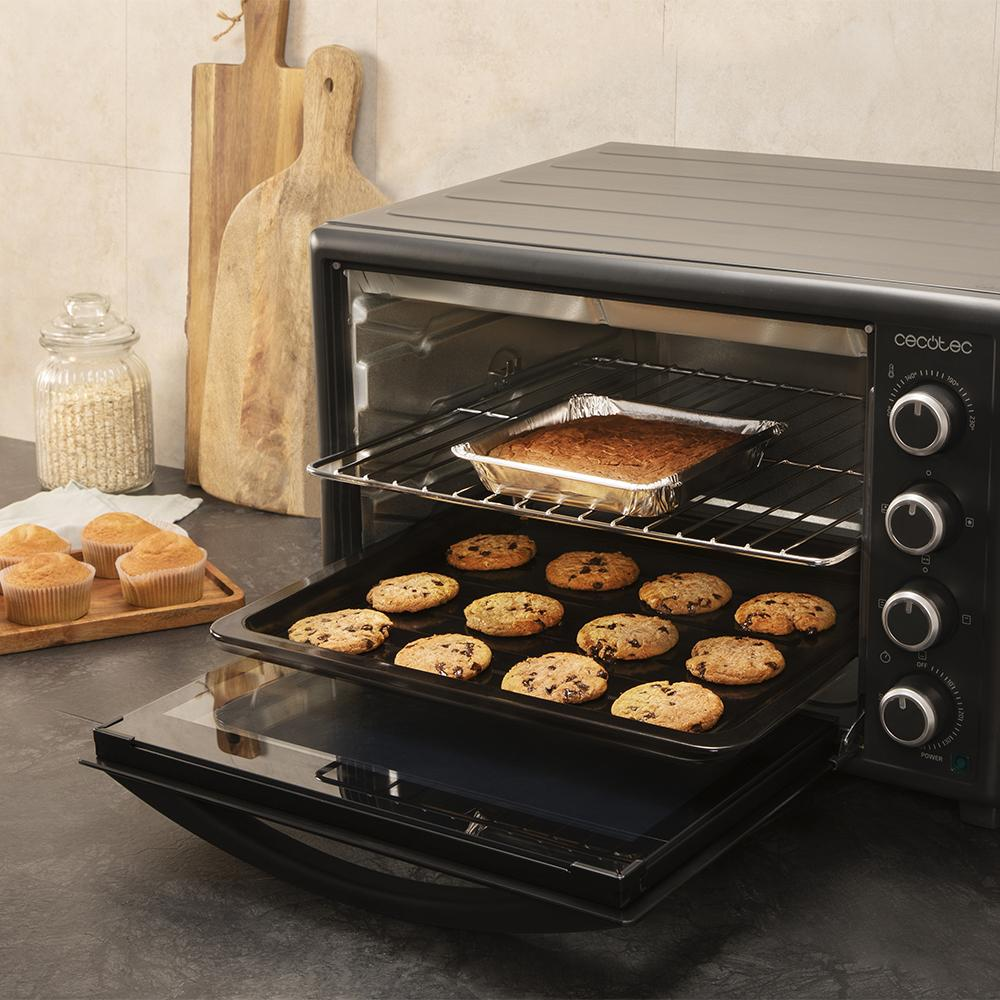 Un horno negro con galletas en una bandeja y un pastel en papel de aluminio dentro. Hay magdalenas en una tabla de madera.