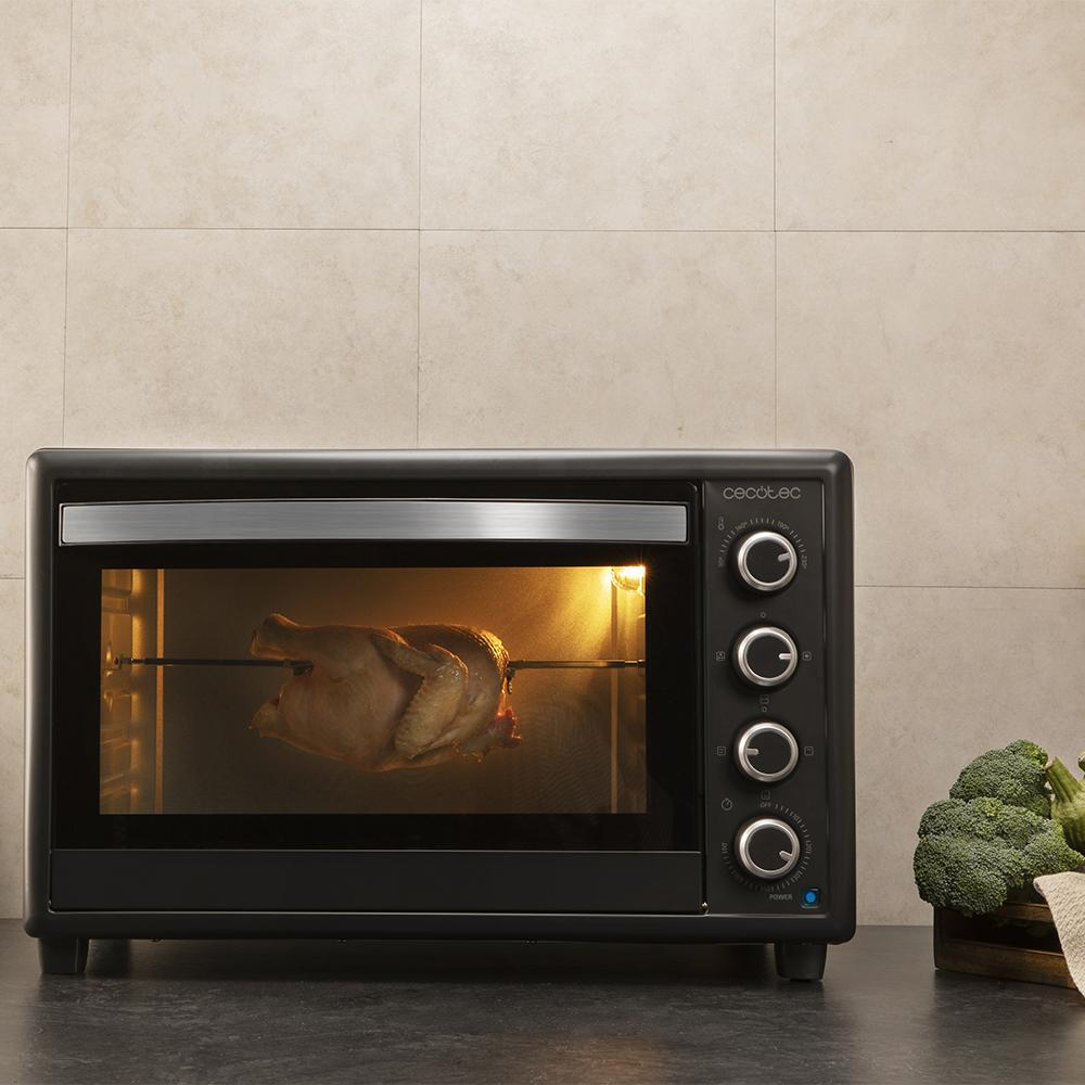 Un pollo entero gira en un asador dentro de un horno negro. Brócoli en una cesta cerca.