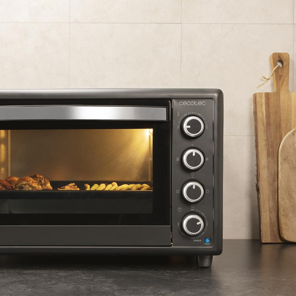Un horno Cecotec negro con comida cocinándose dentro. Cuatro mandos a la derecha, tablas de madera al fondo.