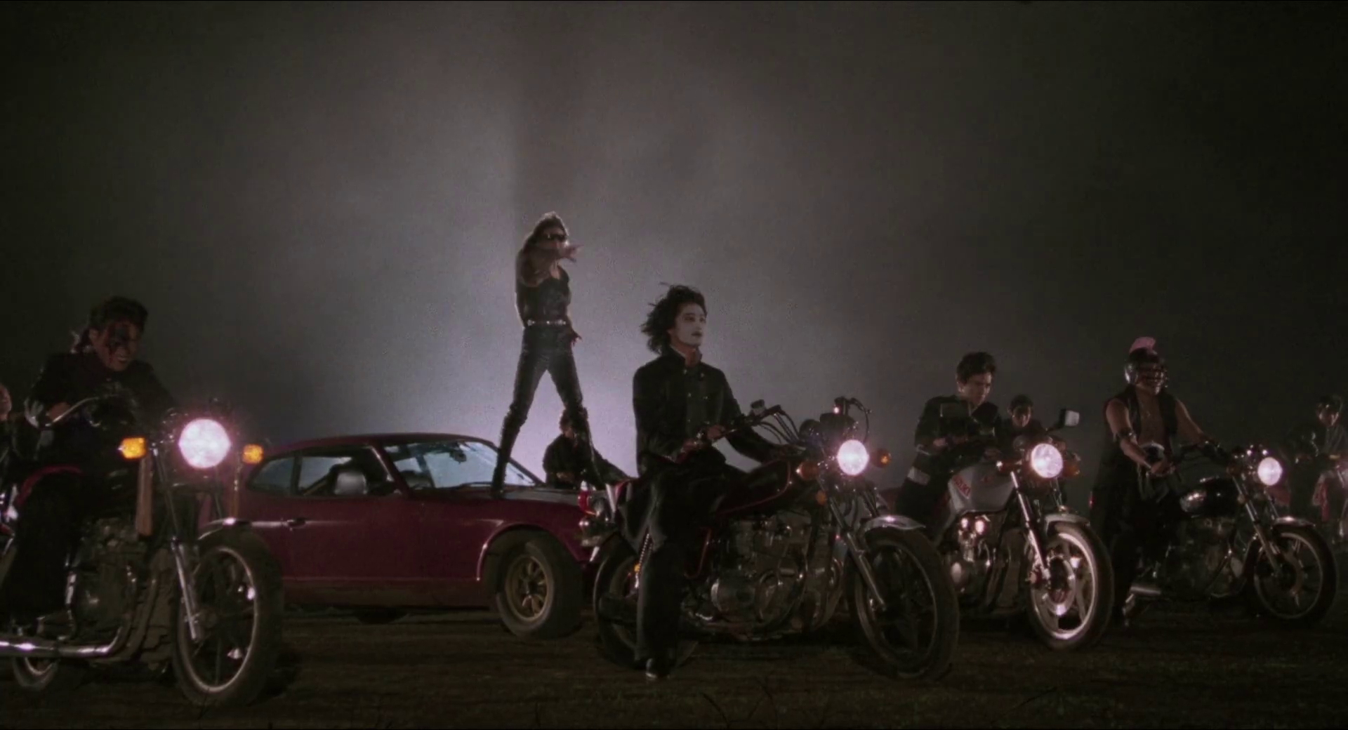 Eine Gruppe von Bikern in dunkler Kleidung steht mit Motorrädern und einem Auto im Nebel.