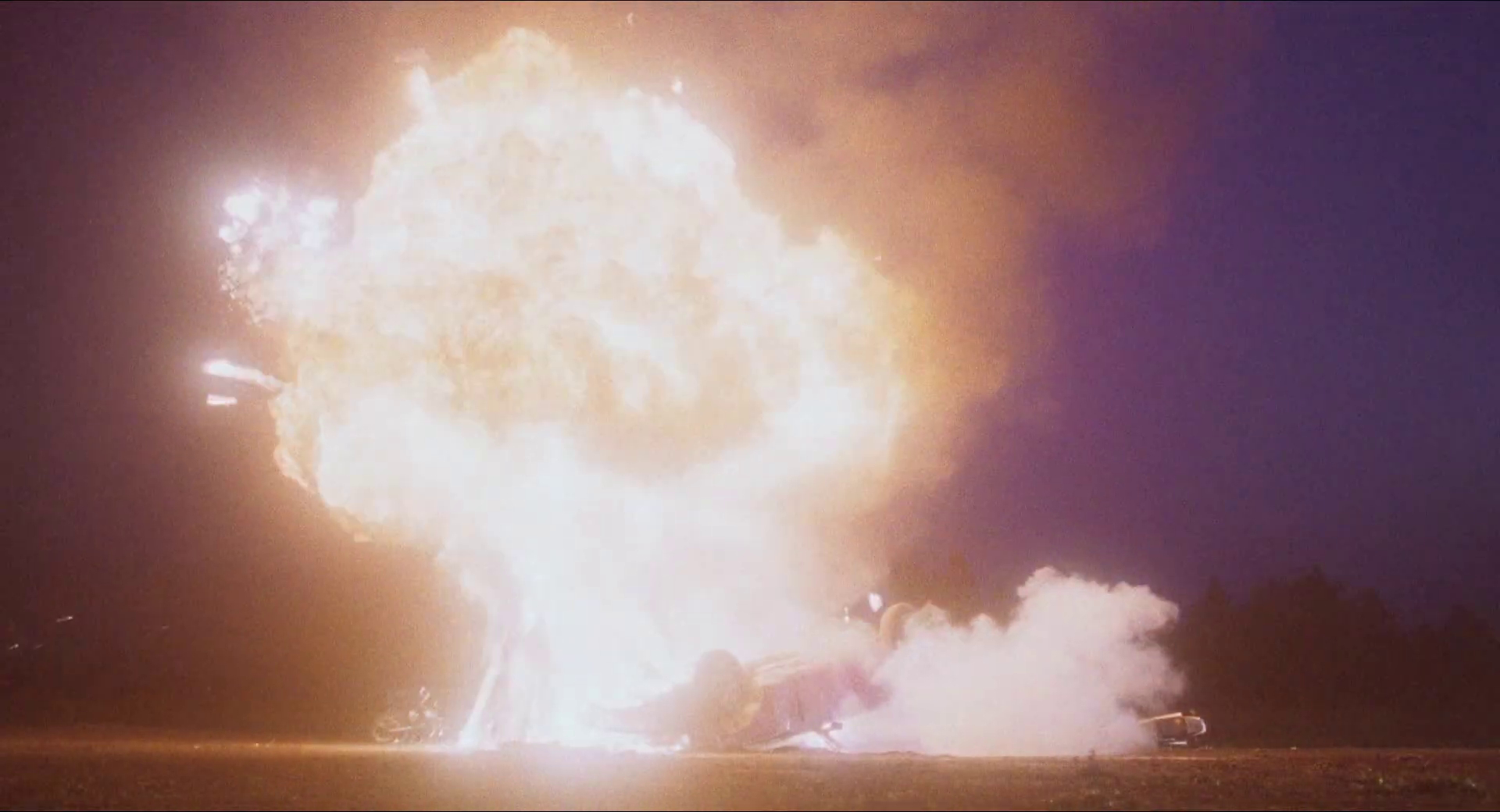 Eine massive Explosion mit Flammen und Rauch ereignet sich über einem umgestürzten Fahrzeug bei Nacht.