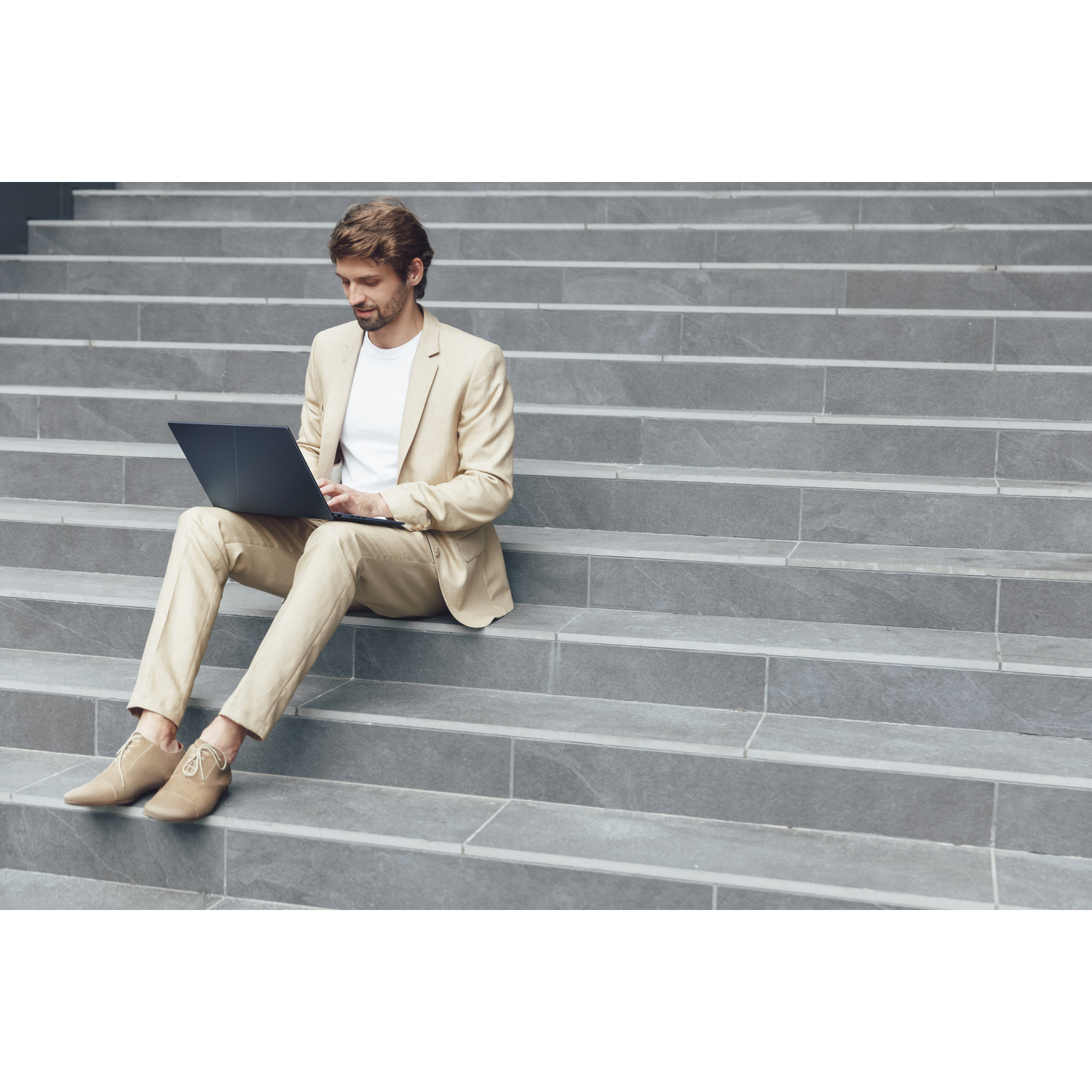 Un uomo in completo beige siede sui gradini di pietra usando un laptop.