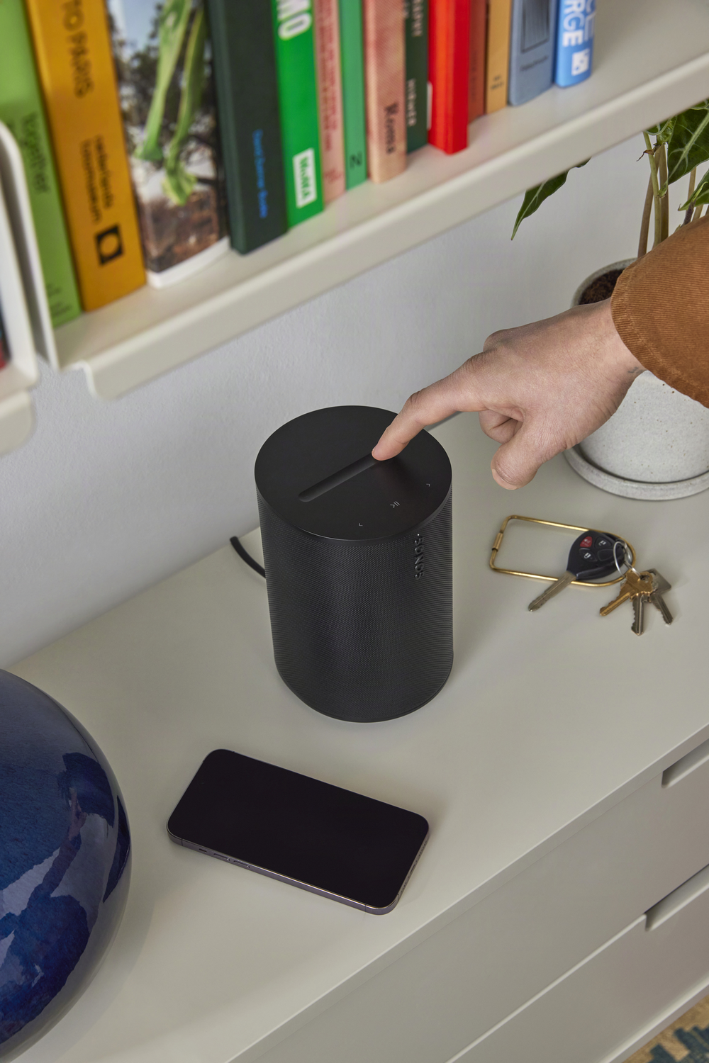 Una mano presiona la parte superior de un altavoz negro. Un teléfono y llaves están cerca.