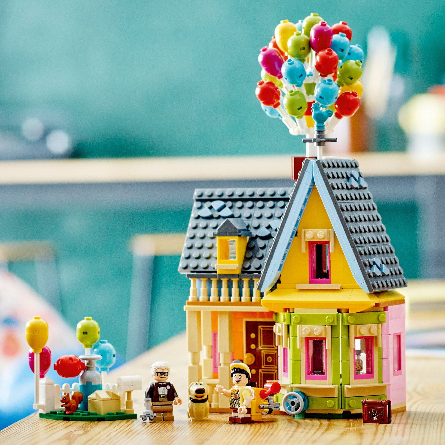 A colorful Lego house with balloons, figures, and a squirrel on a wooden table.