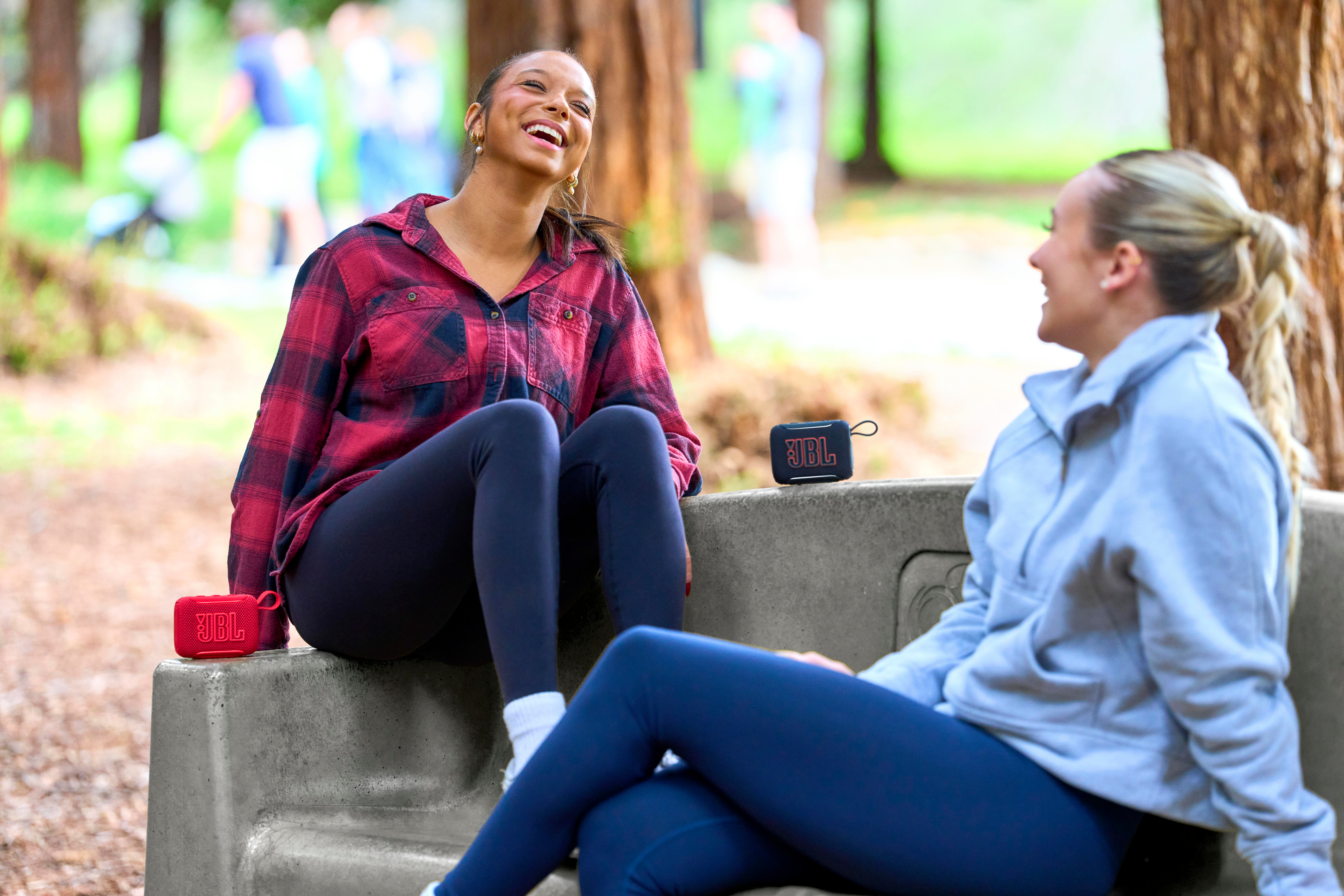 Zwei Frauen auf einer Parkbank mit JBL-Lautsprechern. Eine lacht, trägt ein rotes Flanellhemd.