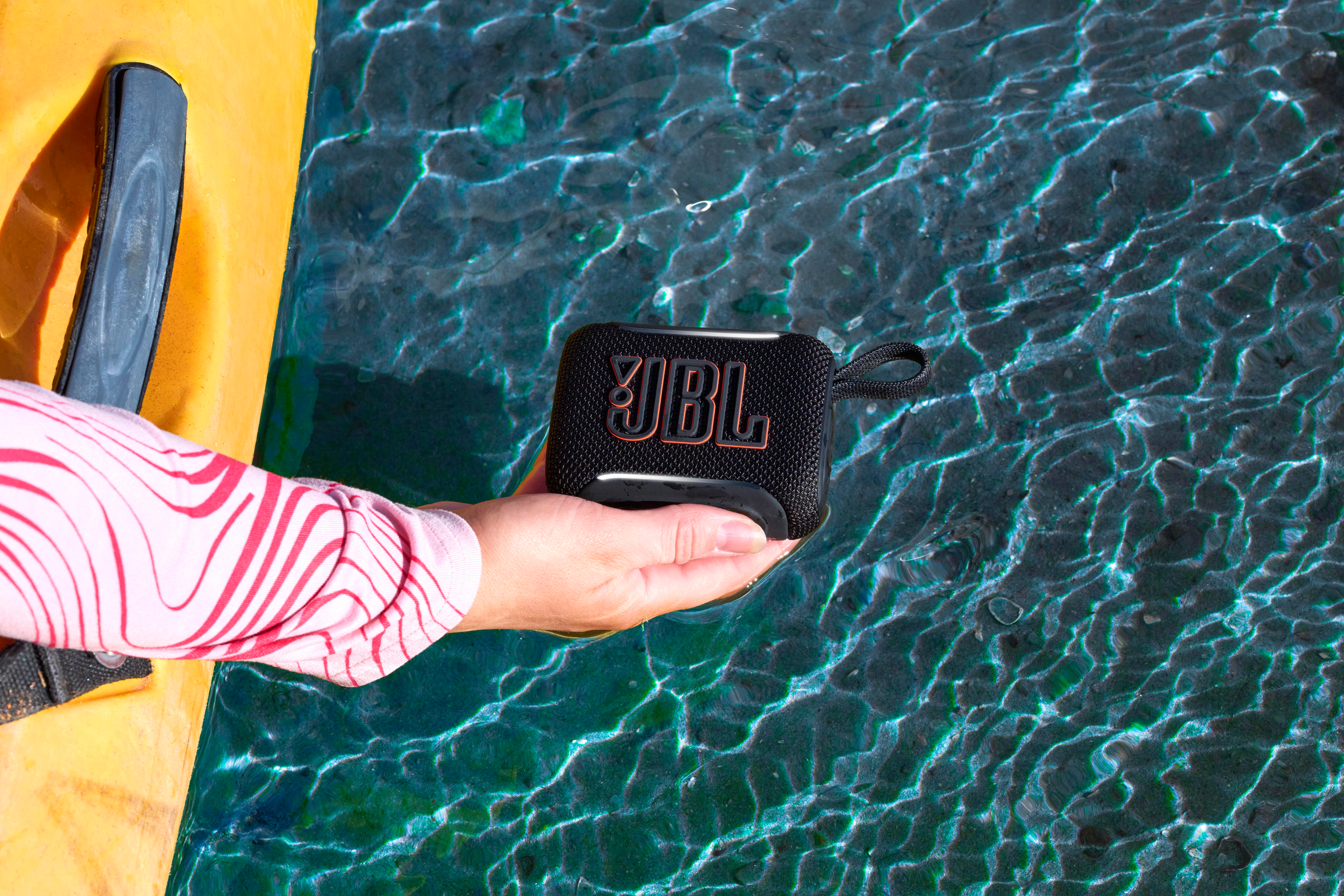 Eine Hand hält einen schwarzen JBL-Lautsprecher über schimmerndem blauem Wasser neben einem gelben Objekt.