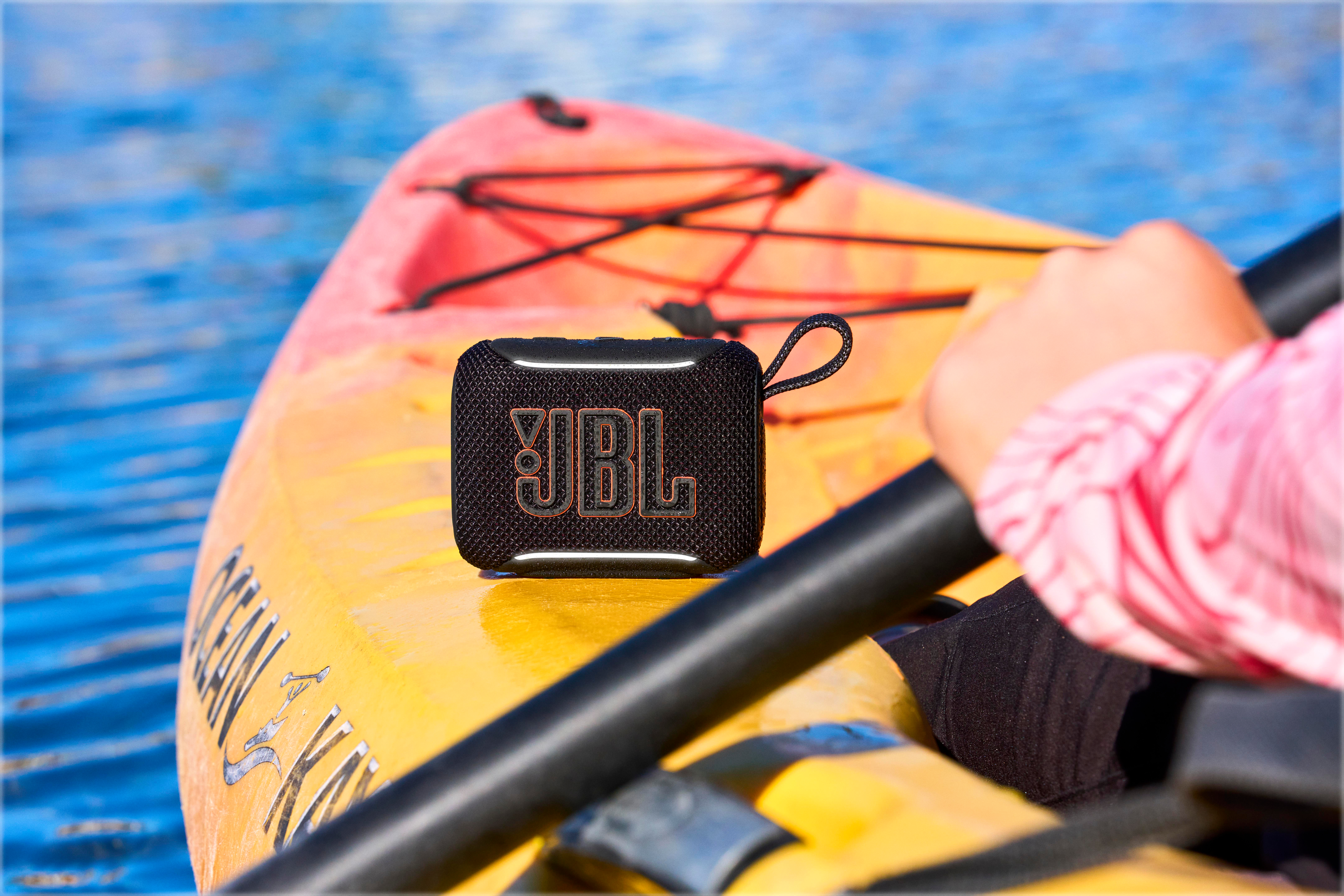 Ein schwarzer JBL-Lautsprecher sitzt auf einem gelben Kajak auf blauem Wasser. Ein Paddel ist sichtbar.