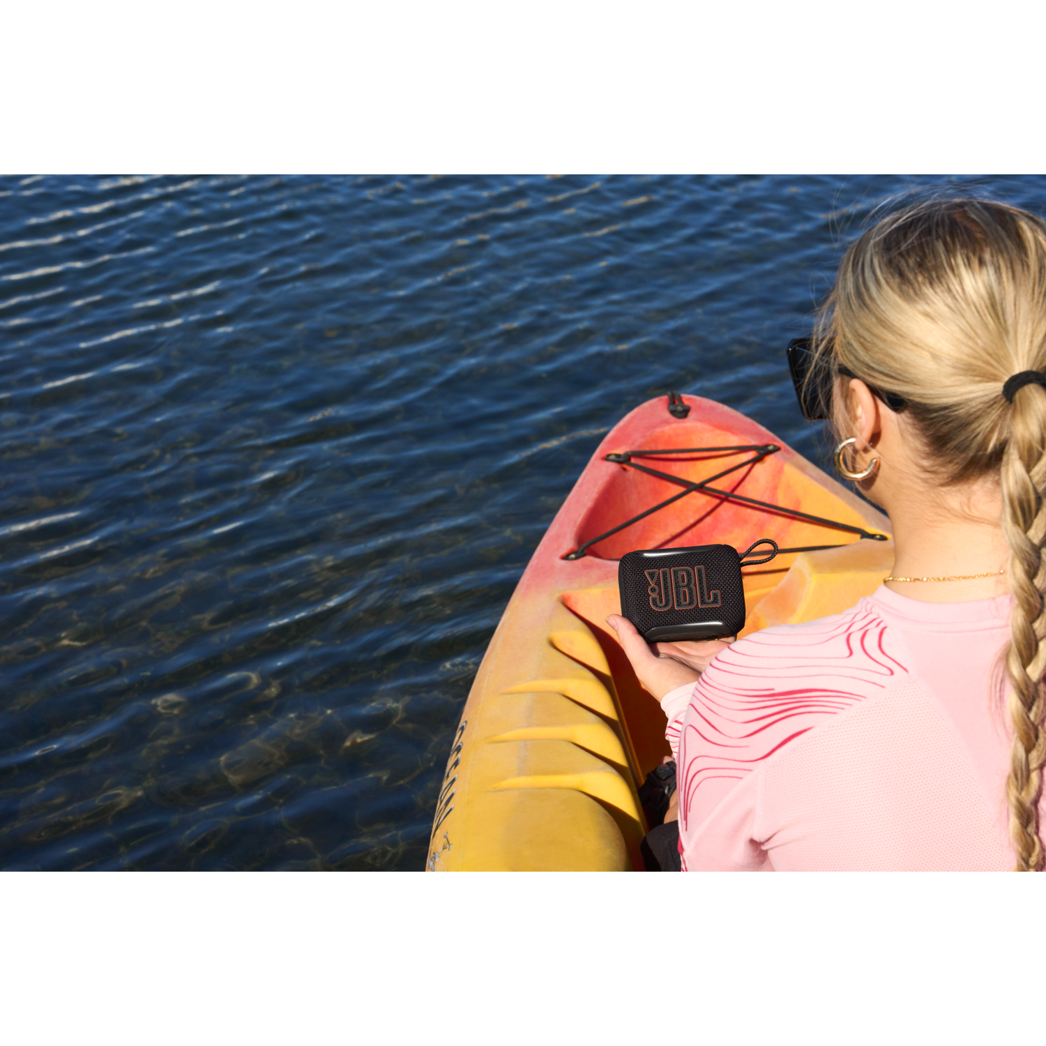 A woman holds a black JBL speaker next to a yellow kayak on blue water.