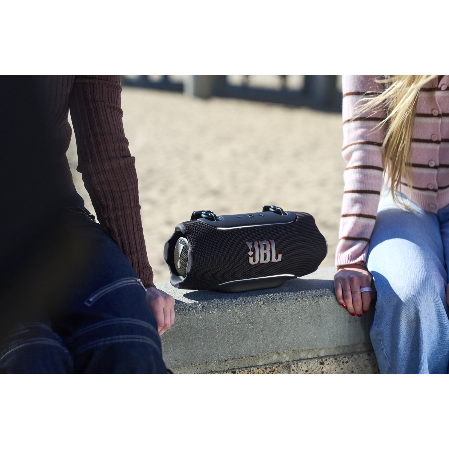 A black JBL speaker sits on a concrete wall. Two people sit beside it.