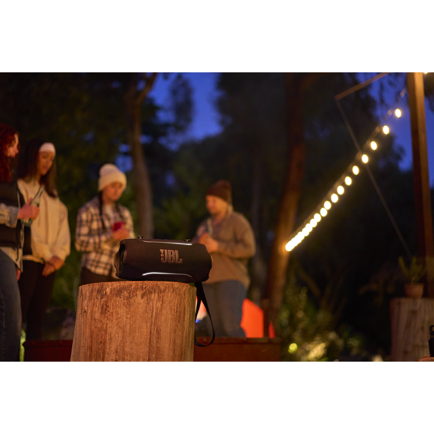 Black JBL speaker on a log, people in the background at night with string lights.