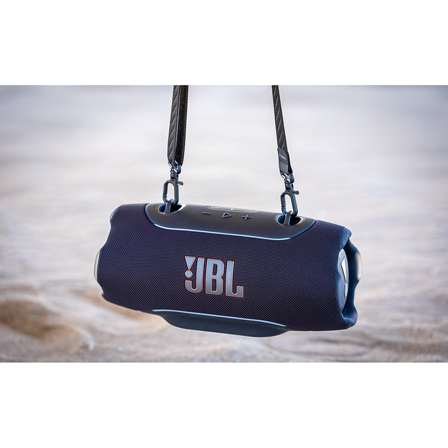 Een blauwe JBL speaker met band hangt boven zand.